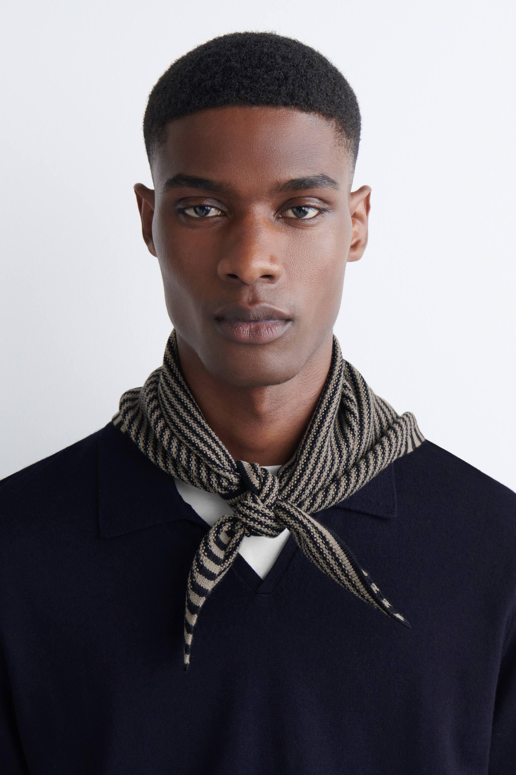 Vezi imaginea mai mare: Un fular tricotat este legat în jurul gâtului, vizibil central din față. Este modelat cu dungi alternante de culoare închisă și bej și este stilizat peste un tricou tricotat de tip polo, de culoare bleumarin închis.