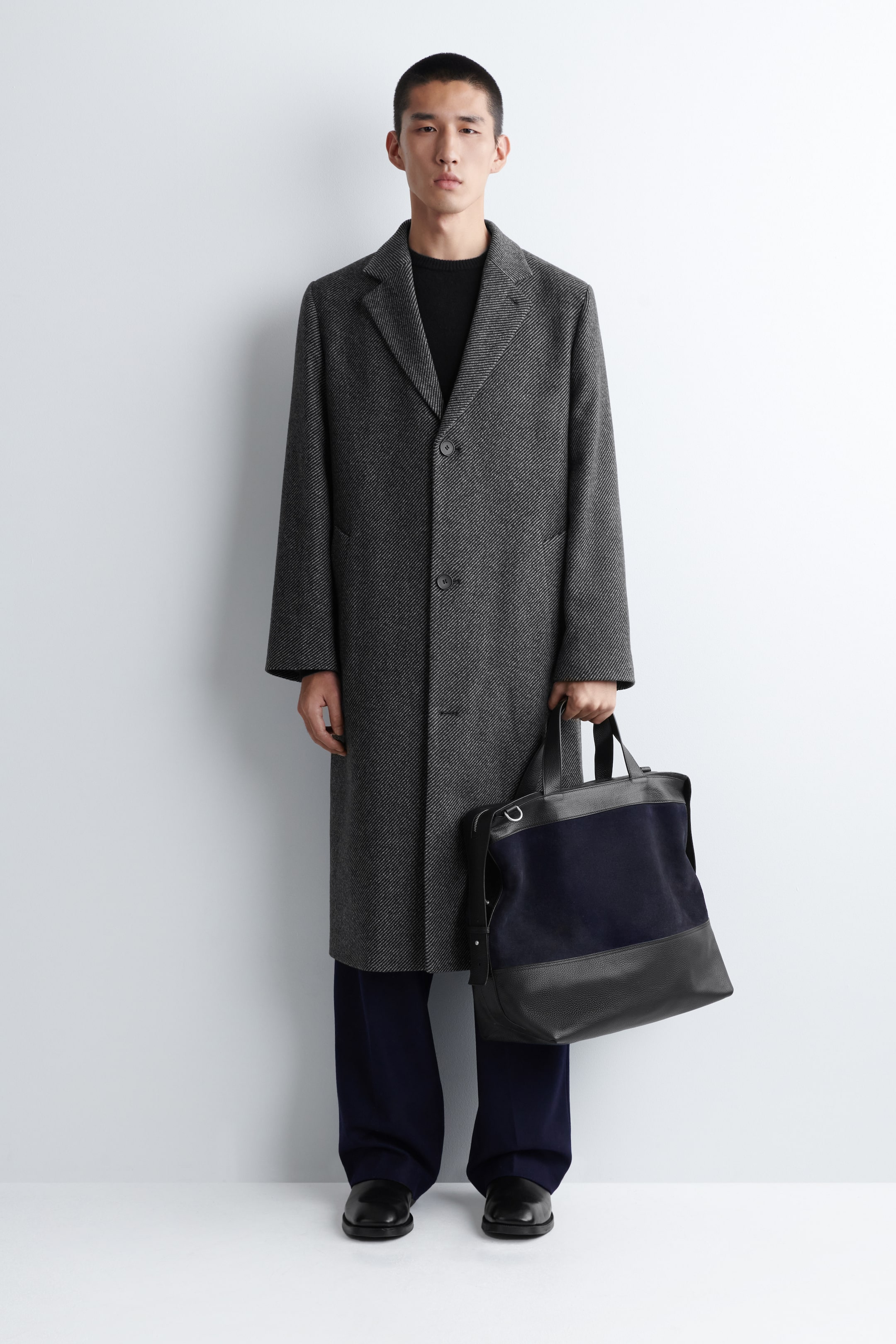 Visualizza immagine più grande: Un cappotto grigio a spina di pesce viene presentato, caratterizzato da revers a dente e un davanti monopetto con tre bottoni visibili. Il lungo cappotto presenta una vestibilità dritta e rilassata ed è stratificato su un maglione nero con scollo a girocollo.