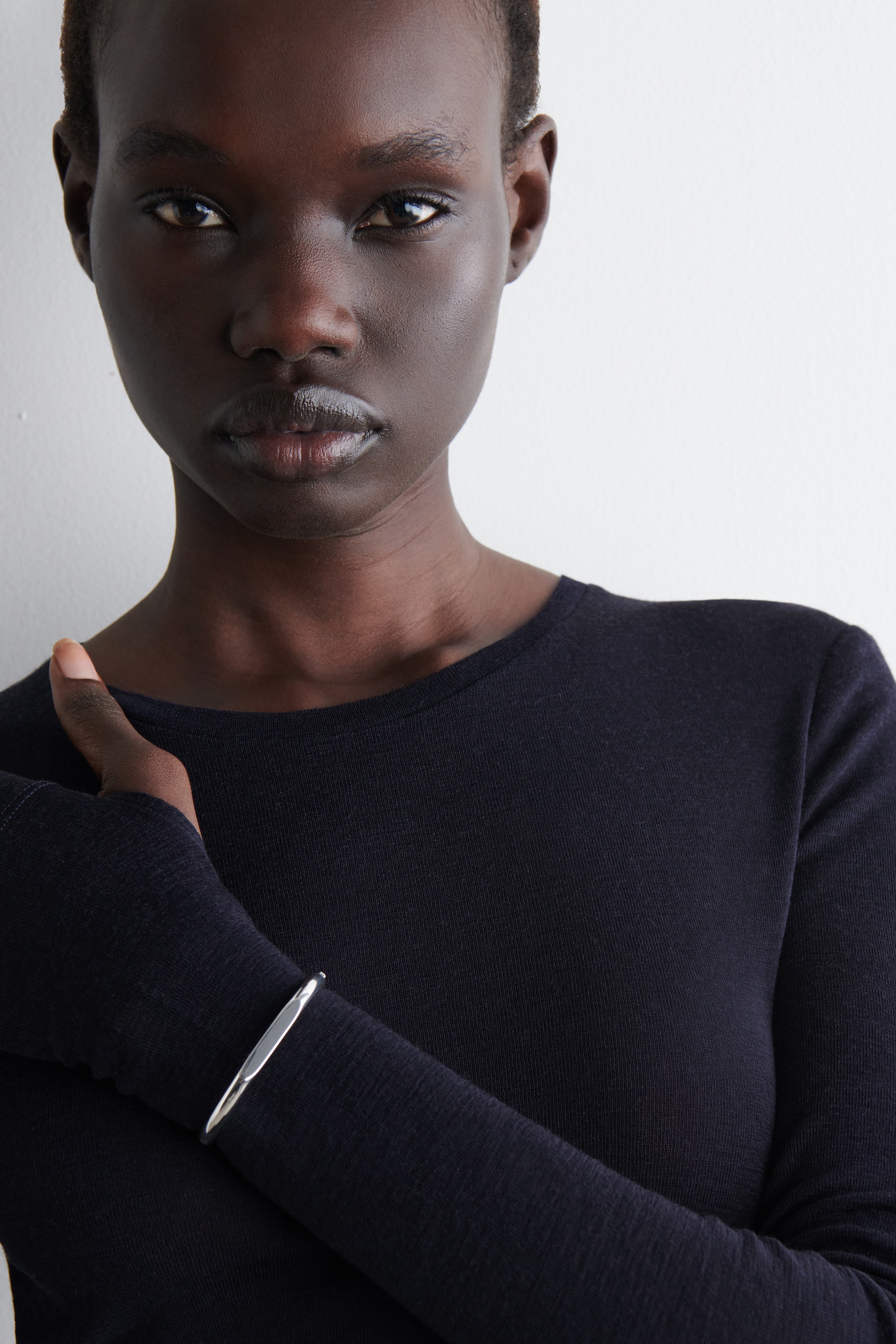 Ver imagen más grande: Se presenta un top oscuro, ajustado y de manga larga, con cuello redondo y mangas extendidas con puños con agujeros para los pulgares. Un brazalete de tono plateado se lleva en la muñeca izquierda sobre la manga.