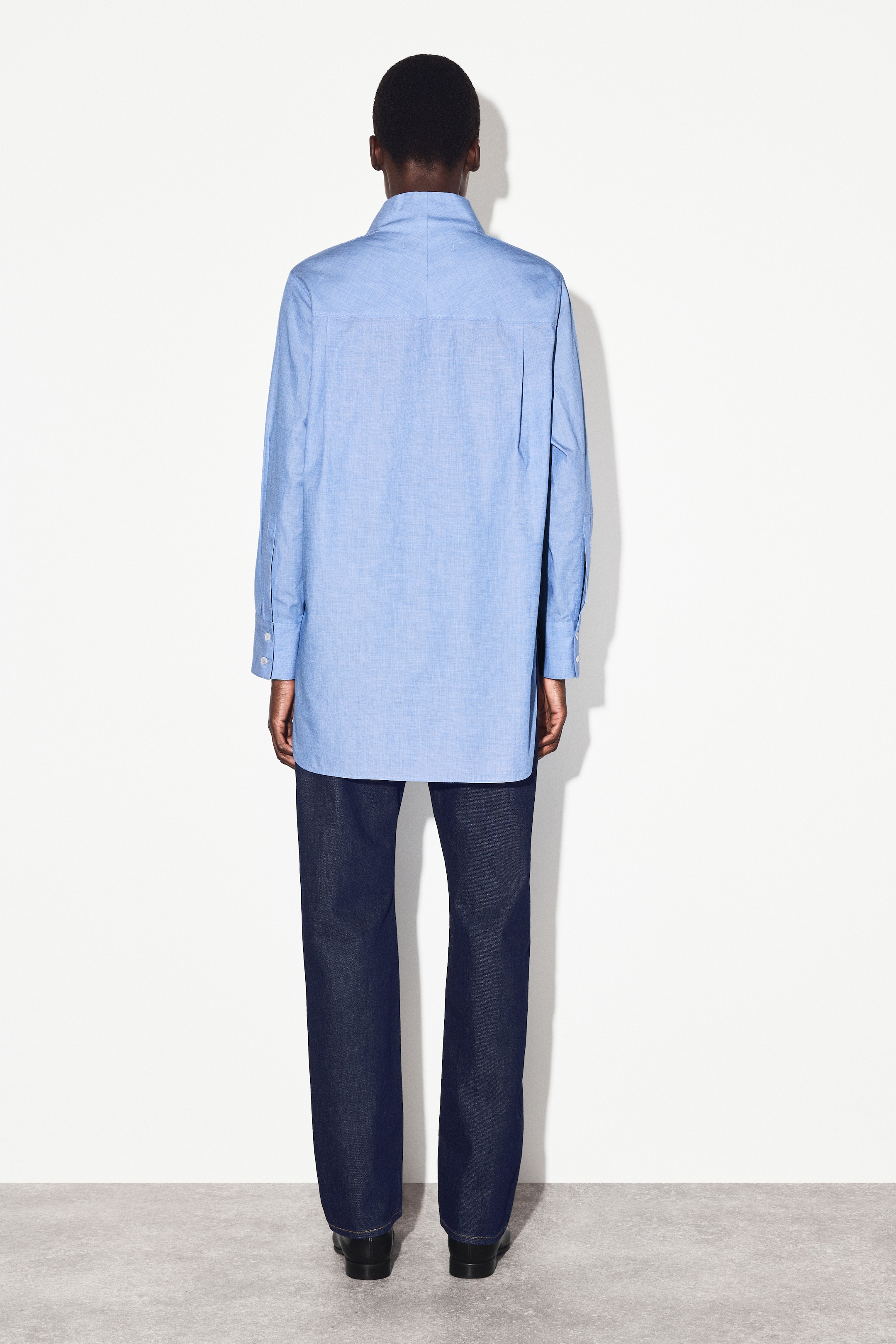 Ingrandisci l'immagine: Una camicia chambray azzurro chiaro è indossata, vista da dietro. Questa camicia oversize con bottoni presenta un colletto a punta, un giogo posteriore, maniche lunghe con polsini abbottonati e un orlo dritto che cade a lunghezza tunica. È abbinata a jeans indaco scuro a gamba larga e scarpe nere con tacco basso.