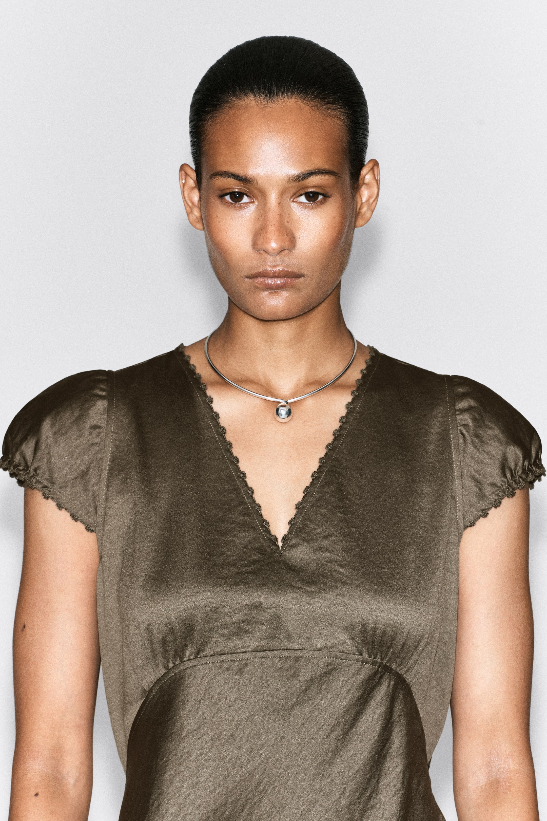 Grotere afbeelding bekijken: Een chokerketting in zilverkleur met een bolvormige hanger wordt gedragen door het model. De ketting rust tegen haar huid, net boven de V-hals van haar donkergroene top.