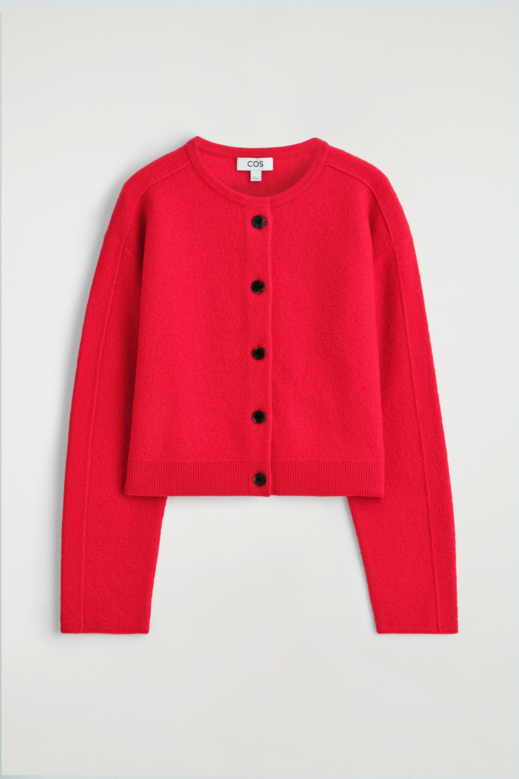 Agrandir l'image: Un cardigan ras du cou rouge se présente à l'avant, avec une laine mérinos bouillie texturée. Il est conçu avec une forme arrondie, des coutures d'épaules superposées, des fermetures à boutons noirs, et des poignets et un ourlet côtelés.