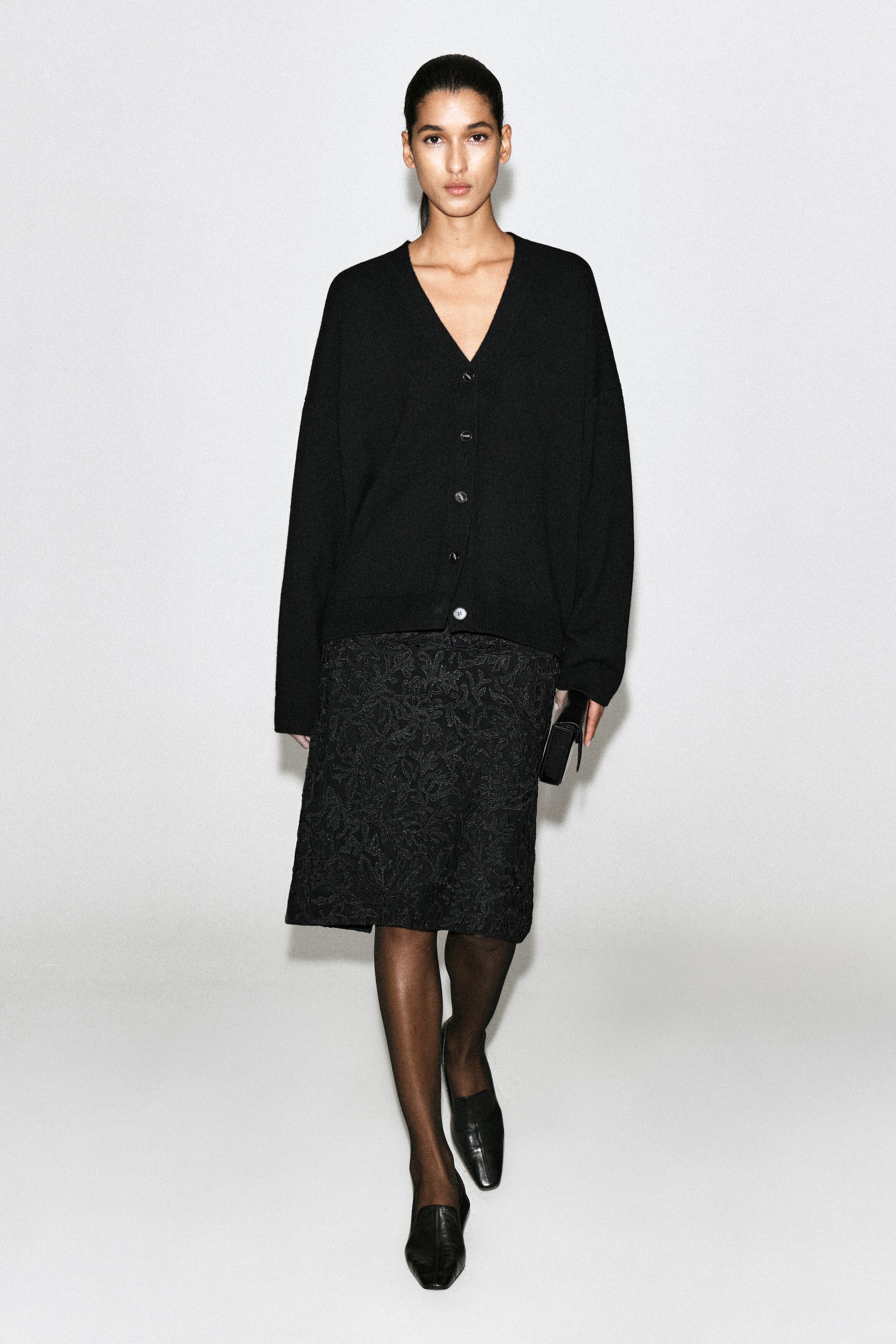 Questo cardigan nero dalla vestibilità rilassata, con scollo a V, è indossato frontalmente e presenta chiusura con bottoni, spalle scese e bordi a coste. È abbinato a una gonna nera al ginocchio con texture, collant neri trasparenti e mocassini neri in pelle.