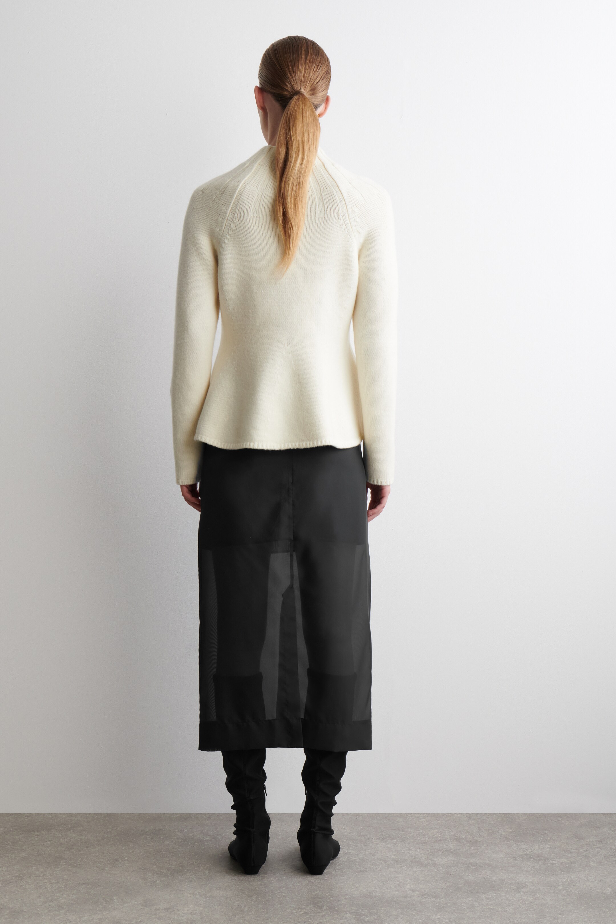 View larger image: A cream sweater is being presented from the back, featuring a high neckline and subtle knit detailing radiating from the collar over the shoulders, with its hem gently flaring. It is layered over a black midi skirt that is showcasing a sheer outer fabric with opaque vertical panels, extending to the calves, and being worn with black, soft boots.