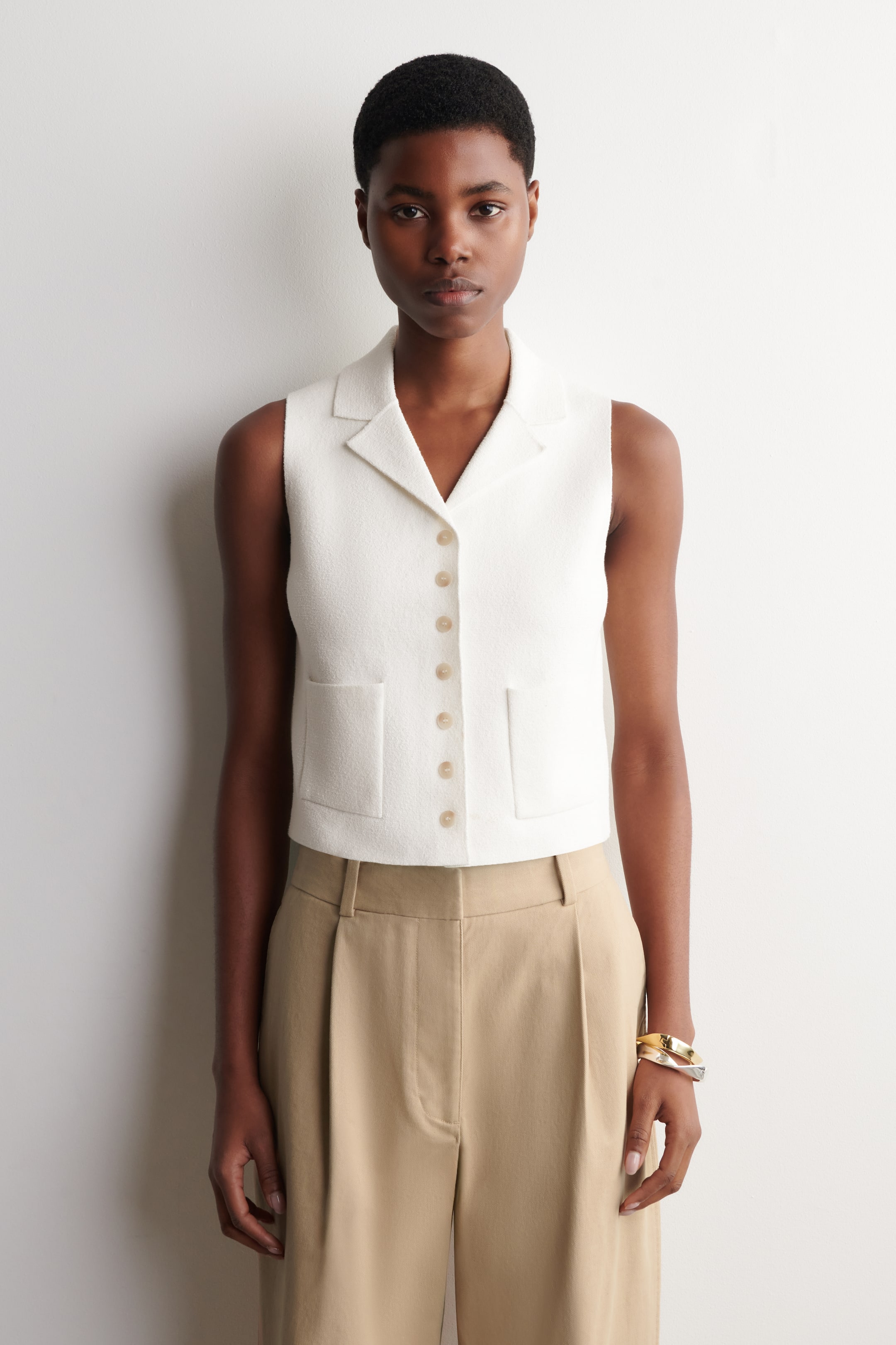 הצגת תמונה גדולה יותר: הדוגמנית לובשת וסט סרוג מבוקלה בצבע לבן, הכולל צווארון נופש, סגירת כפתורים מנוגדת ושני כיסי טלאי קדמיים. הווסט מותאם למכנסיים מכווצים בצבע חום בהיר.