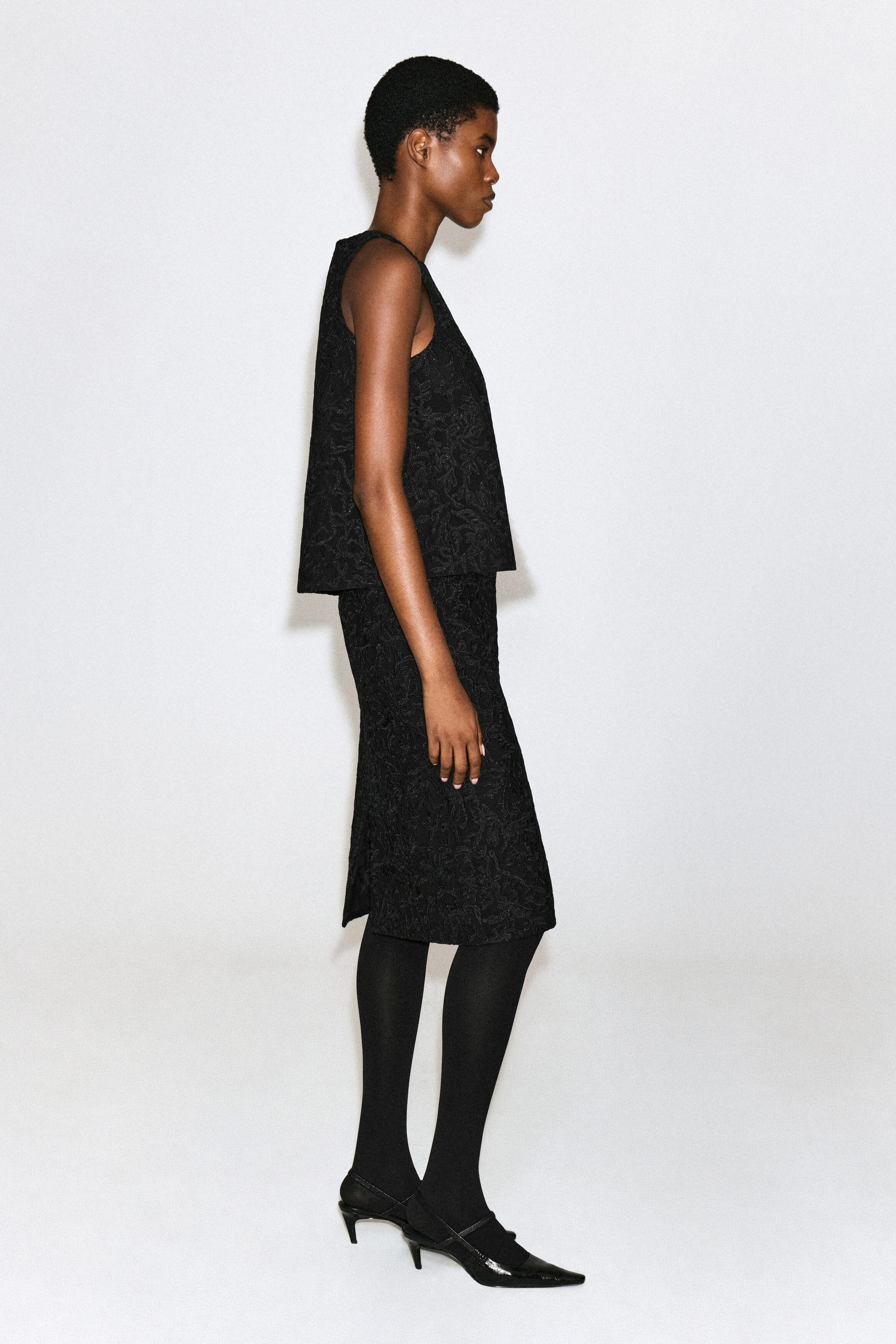 Un haut sans manches noir orné d’un motif floral en dentelle texturée est porté par le mannequin de profil. Le haut à coupe décontractée est agencé avec une jupe texturée assortie, des collants noirs et des escarpins noirs à bout pointu.