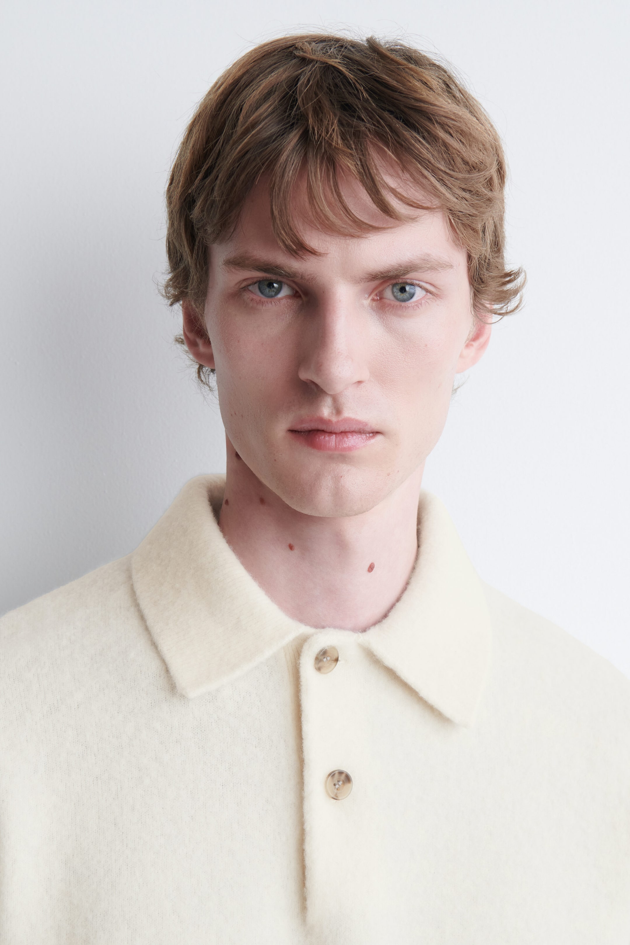 The model is wearing a cream-colored brushed wool polo shirt, showcasing a soft, textured fabric. The shirt features a classic pointed collar and a front placket with two light-colored buttons.