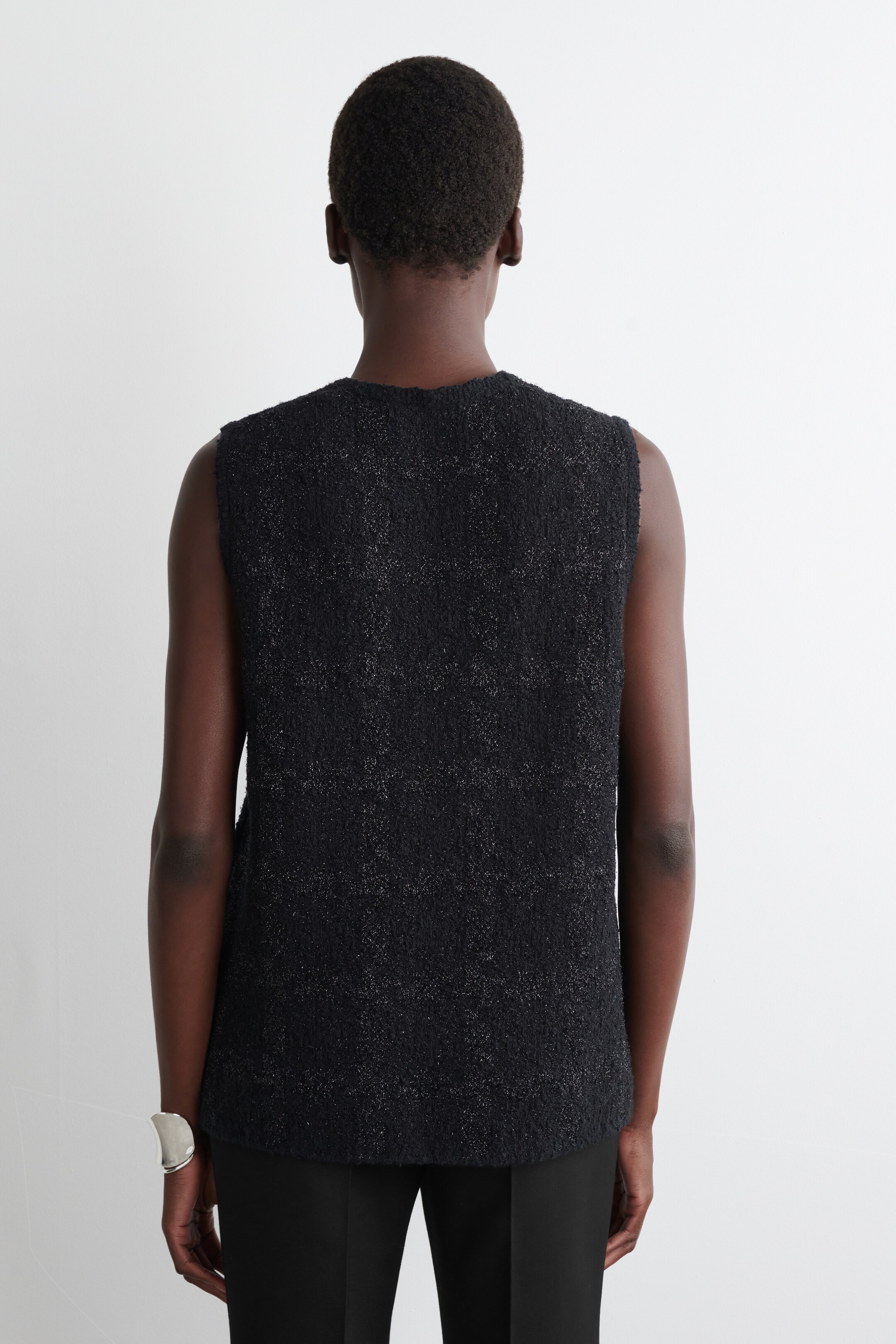Agrandir l'image: Un haut sans manches noir est présenté de dos. Il présente un tissu texturé parsemé de fibres légères et un col rond, tombant droit jusqu'à un ourlet simple tout en étant porté sur un pantalon noir.