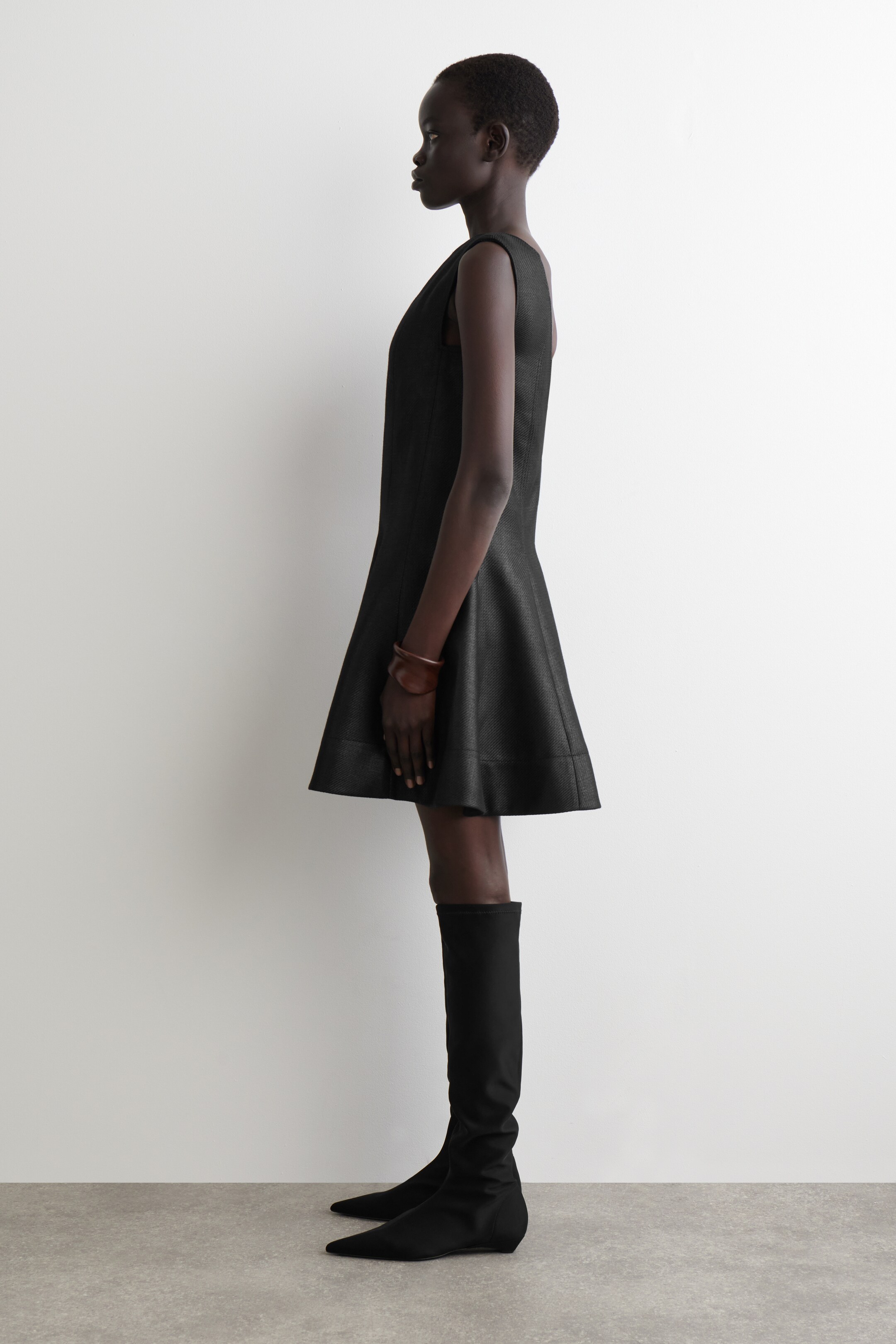 View larger image: A black sleeveless dress with a textured weave, featuring an A-line silhouette and a hem ending above the knee, is being presented from a side profile. It is being styled with black knee-high, pointed-toe flat boots and a single brown bracelet on the wrist.