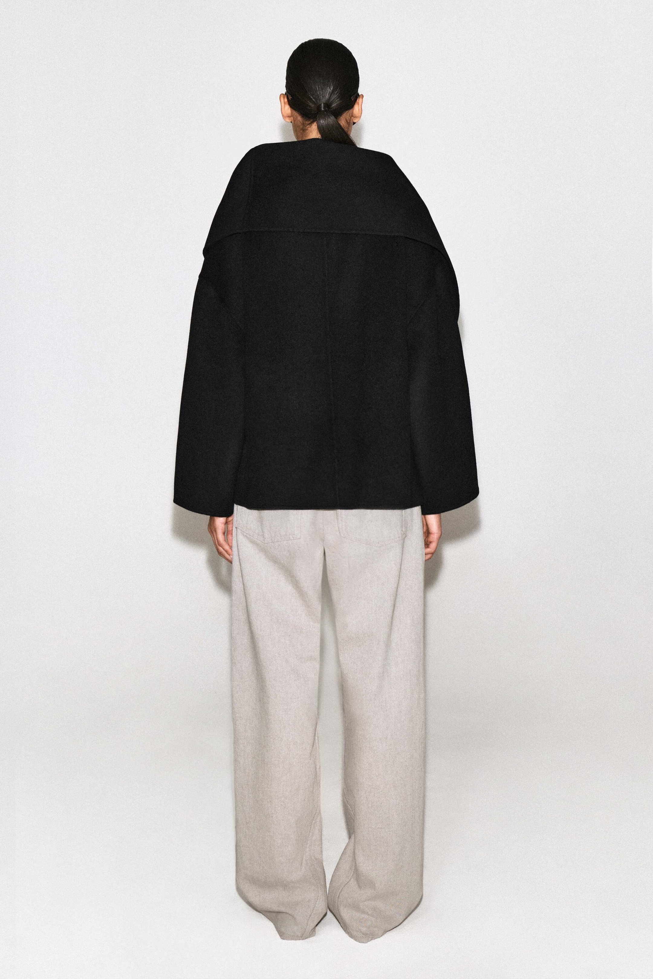 The double-faced wool-blend scarf jacket features a relaxed, oversized silhouette in black, shown from the back to highlight its wide, draped collar resting across the shoulders. The jacket has straight, wide-cut sleeves and falls to the mid-hip, styled with light-colored wide-leg trousers.