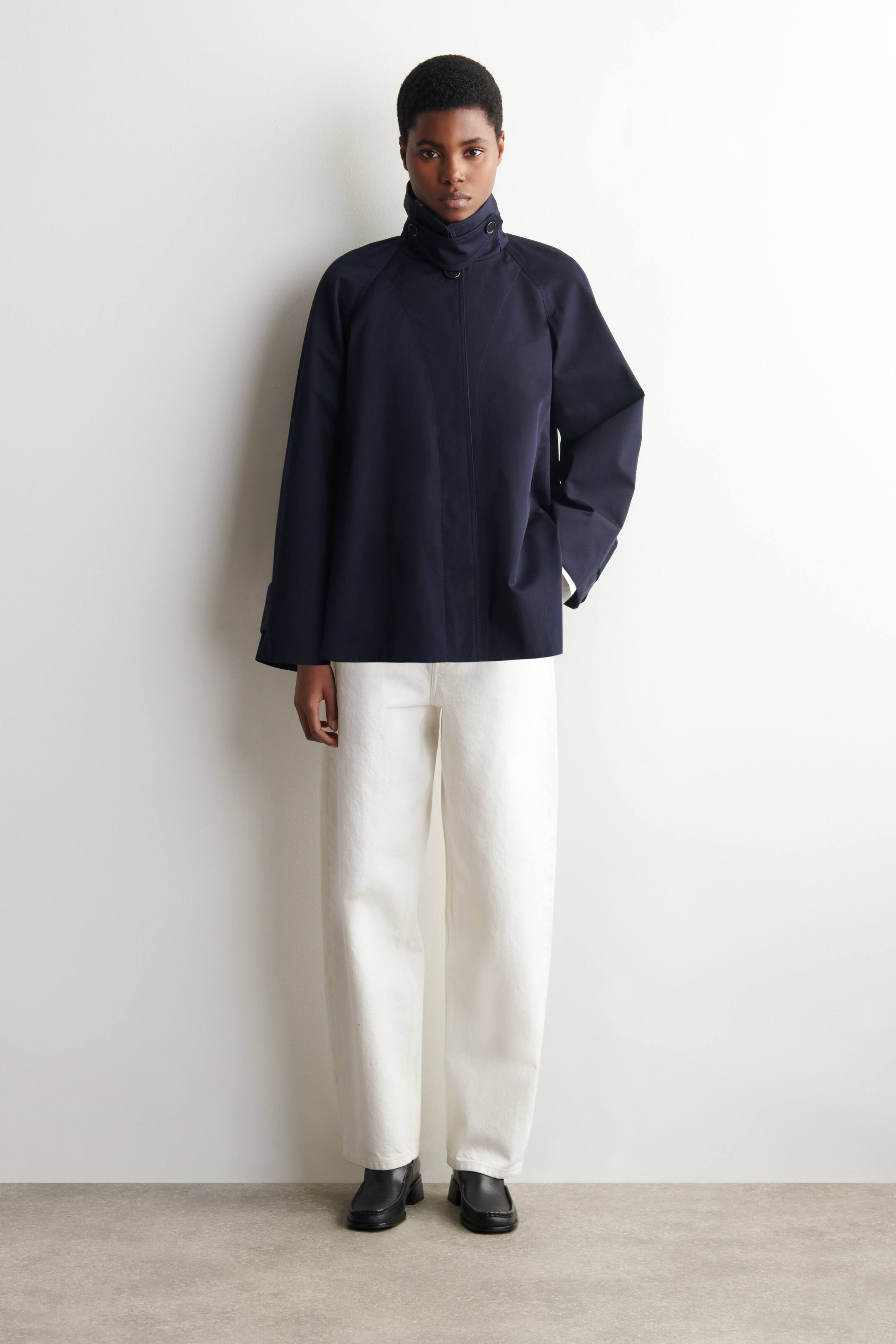 Agrandir l'image: La mannequin porte une veste trapèze bleu marine avec une fermeture à boutons. Son col est stylé en col cheminée haut, fixé avec des boutons visibles.