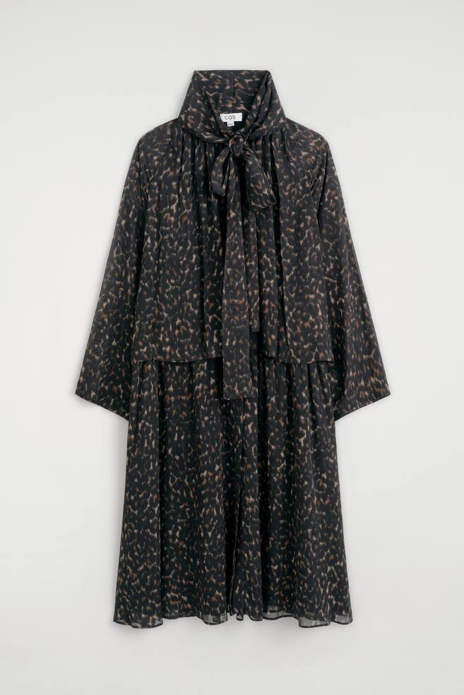 The leopard-print fluid scarf-detail dress is hanging in a front view, displaying a high collar with a self-tie scarf detail knotted at the neck. It features wide, long sleeves, a layered bodice design, and a relaxed, fluid silhouette with an all-over brown and black leopard pattern. - 1