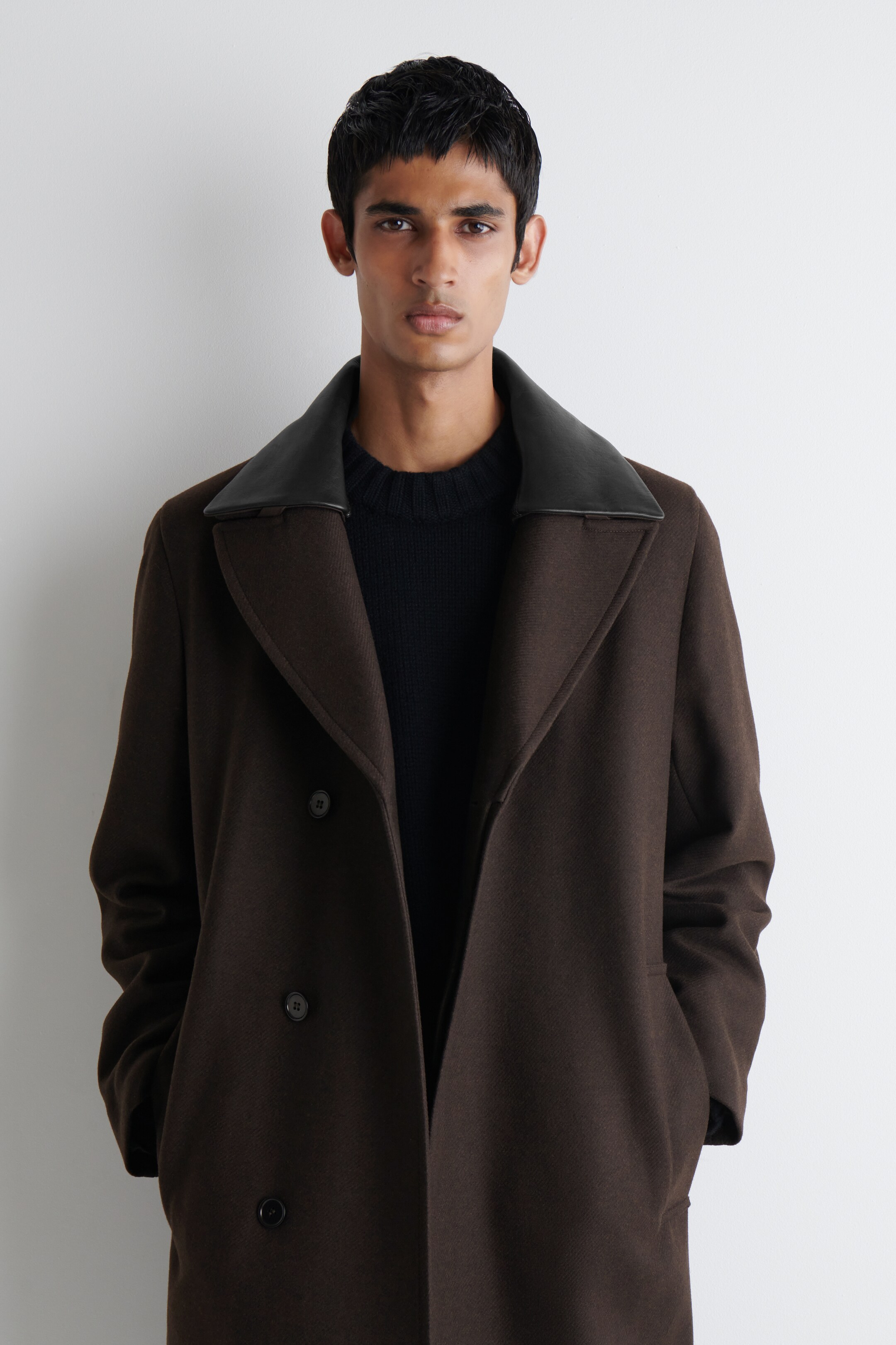 Visualizza immagine più grande: Un cappotto marrone doppiopetto è indossato aperto, rivelando un maglione nero, e presenta ampi revers a lancia, un colletto in pelle nera a contrasto e tasche a filetto visibili.