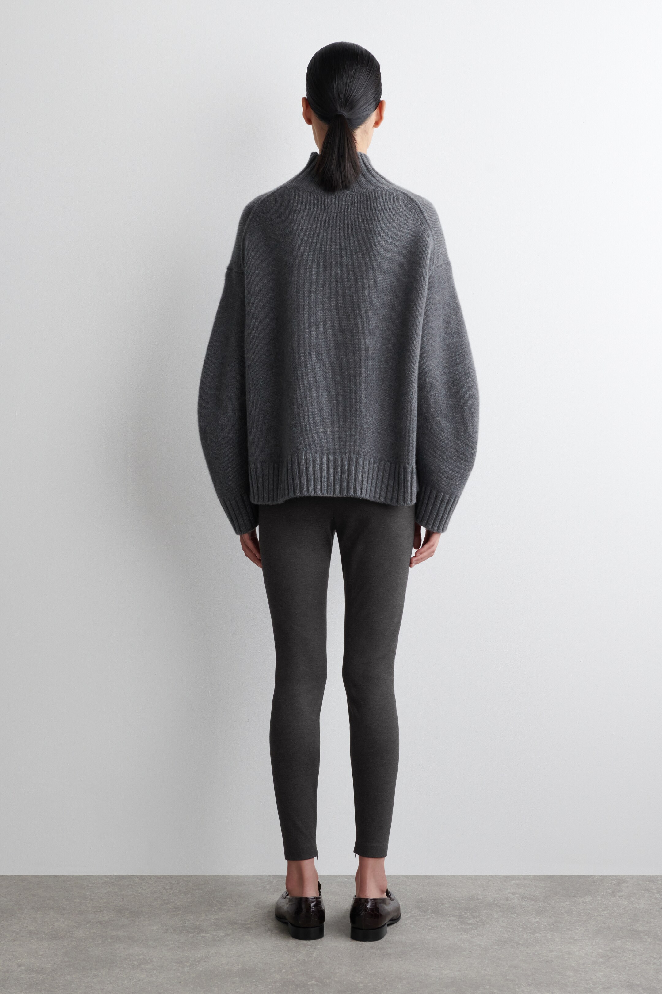 Grotere afbeelding bekijken: Een donkergrijze oversized trui wordt gepresenteerd vanaf de achterkant, met een hoge nek, volumineuze lange mouwen en een geribde zoom. Het wordt gestyled met donkergrijze slim-fit leggings, die enkellengte ritsen hebben, en donkerbruine croco-embossed loafers met een band.