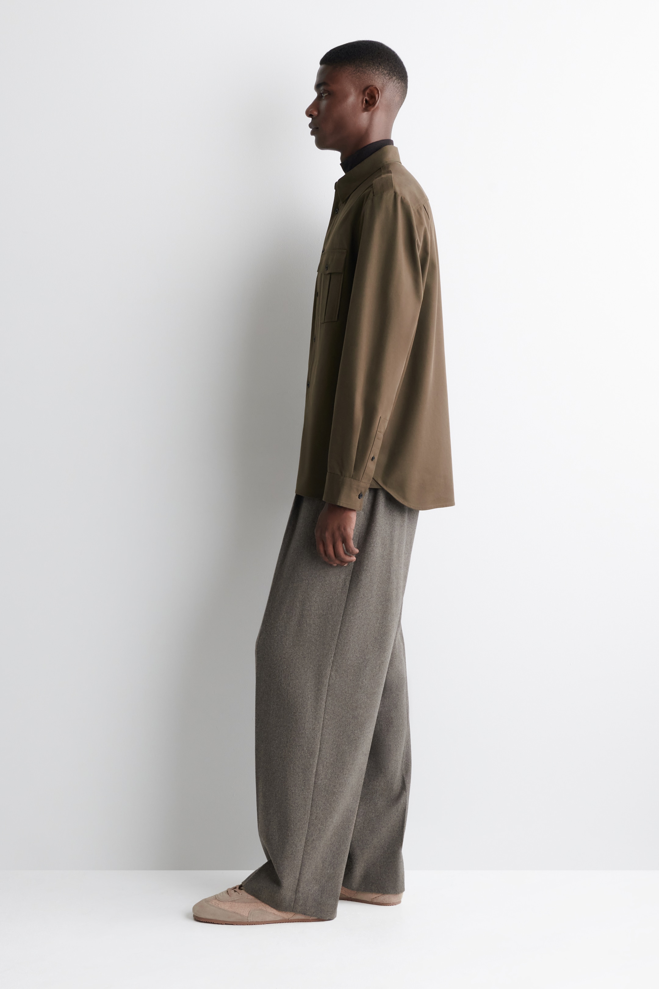 Agrandir l'image: Une surchemise brun olive est présentée de profil, avec deux poches à rabat sur la poitrine et des poignets boutonnés, portée ouverte sur un col roulé noir. Elle est associée à un pantalon ample à jambes larges gris chiné et des baskets beige clair.