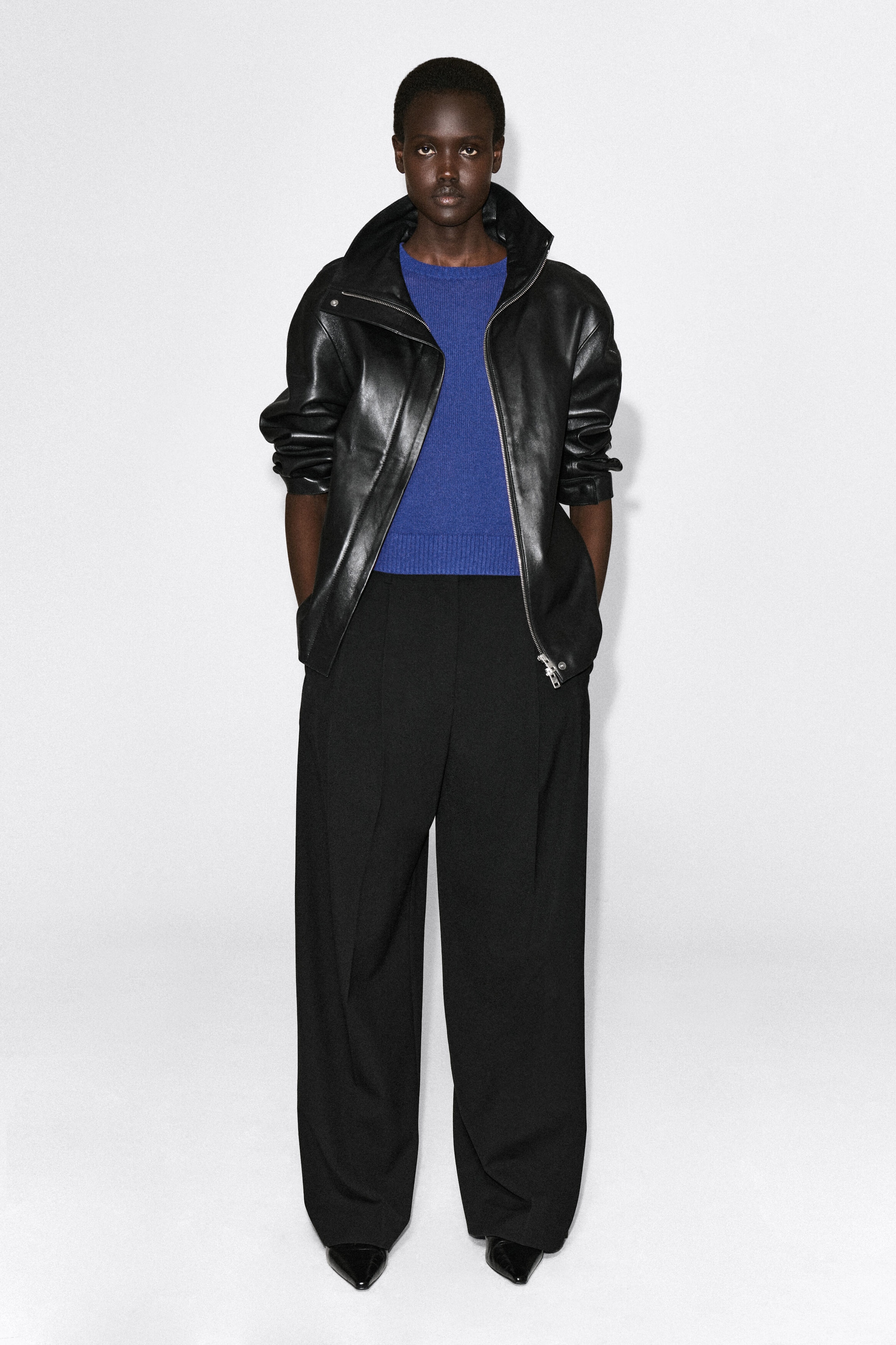 The shrunken knitted linen T-shirt in vibrant blue is shown from the front, highlighting its crew neck, fine knit texture, and ribbed hem. It is layered under an open black leather jacket and paired with high-waisted black trousers, showcasing its shrunken, cropped fit.