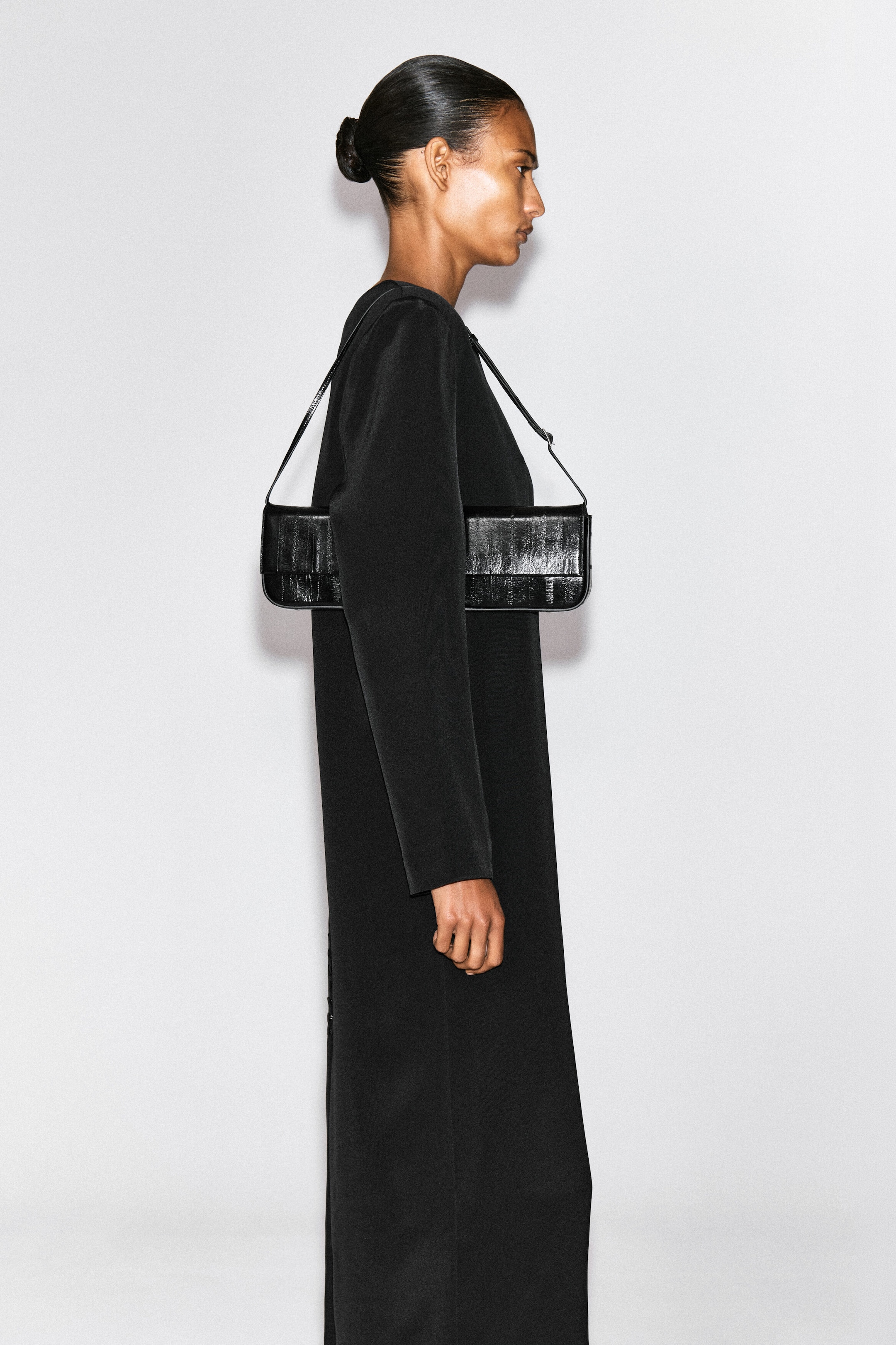 Eine schwarze, längliche rechteckige Schultertasche mit glänzender, segmentierter Struktur wird über der Schulter des Models getragen. Die Tasche verfügt über einen dünnen Riemen und ist über einem langärmeligen schwarzen Kleid gestylt. Eine schwarze, längliche rechteckige Schultertasche mit glänzender, segmentierter Struktur wird über der Schulter des Models getragen. Die Tasche verfügt über einen dünnen Riemen und ist über einem langärmeligen schwarzen Kleid gestylt.