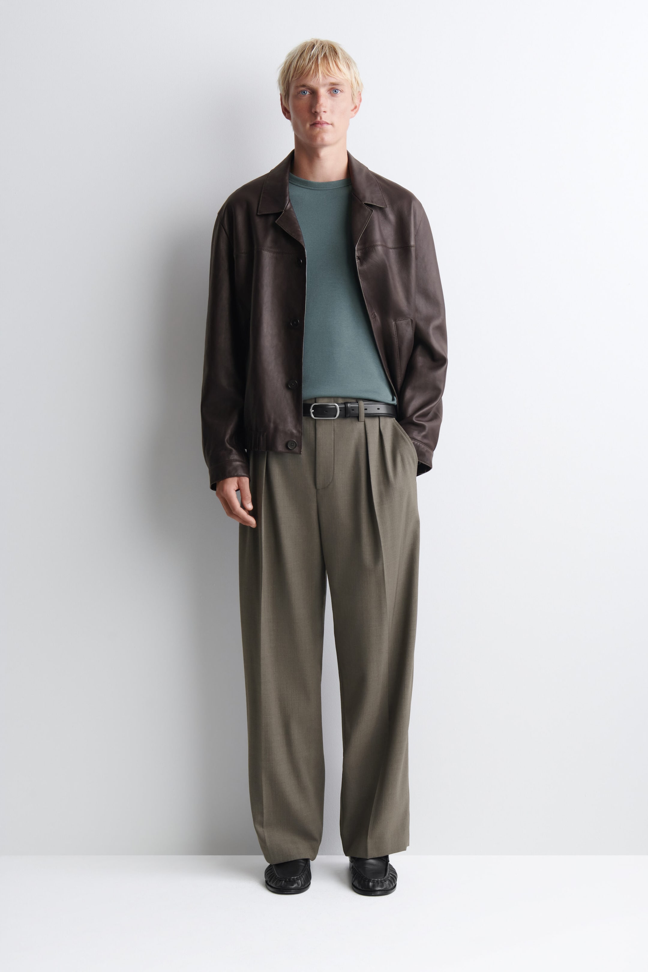 A dark brown leather jacket, shown from the front, features a relaxed fit, a spread collar, front button closures, and side slip pockets. It is styled open over a muted teal crewneck top and olive green wide-leg pleated trousers. A dark brown leather jacket, shown from the front, features a relaxed fit, a spread collar, front button closures, and side slip pockets. It is styled open over a muted teal crewneck top and olive green wide-leg pleated trousers.
