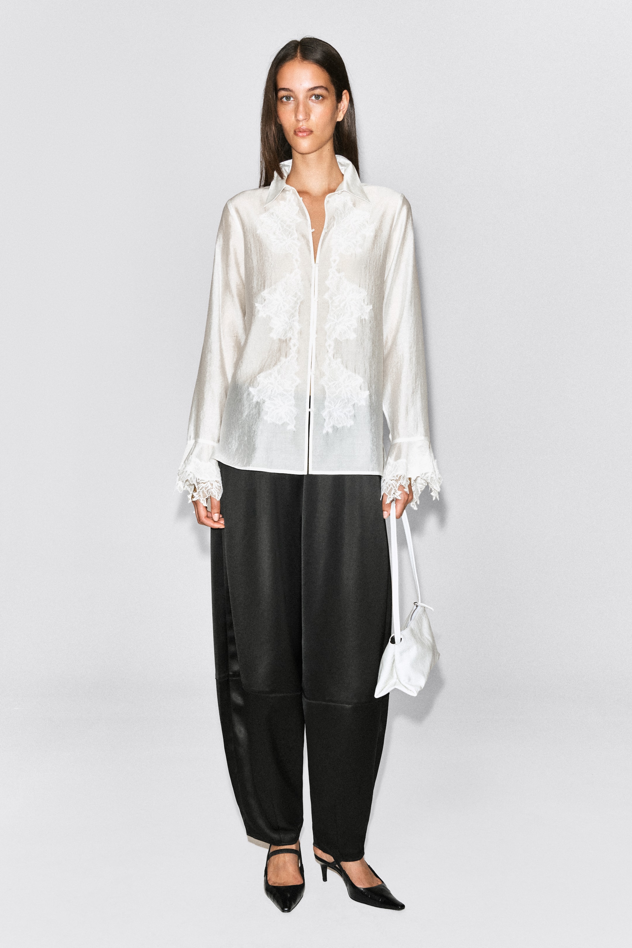 View larger image: Black satin trousers featuring a barrel-leg silhouette and tonal seam detailing are being worn by the model. The trousers are styled with a white long-sleeved blouse with lace embellishments, black pointed-toe heels, and a small white handbag that the model is holding.