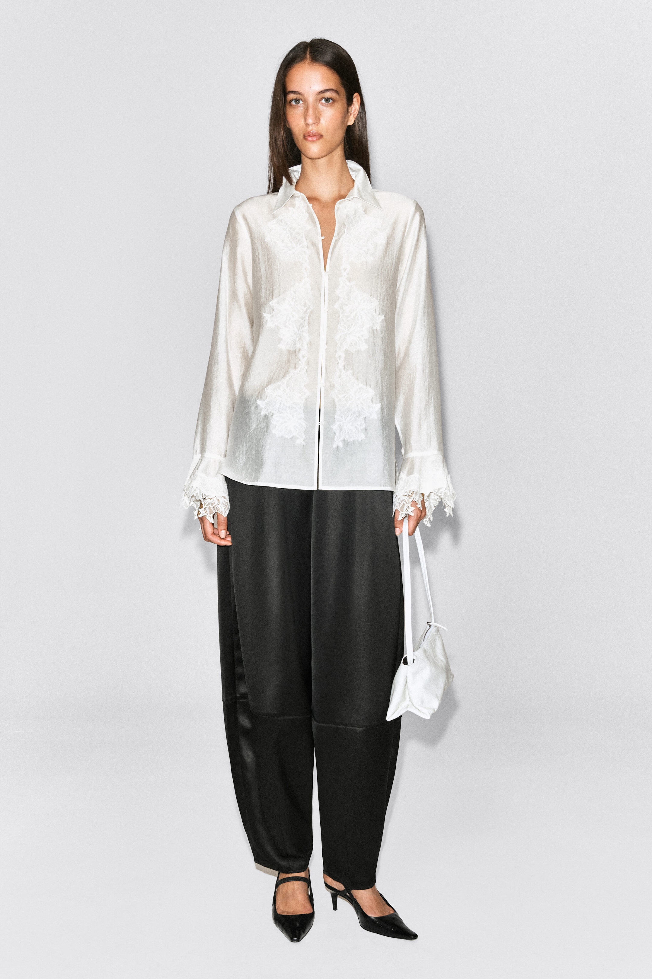 Black satin trousers featuring a barrel-leg silhouette and tonal seam detailing are worn by the model. The trousers are styled with a white long-sleeved blouse with lace embellishments, black pointed-toe heels, and a small white handbag held by the model. Black satin trousers featuring a barrel-leg silhouette and tonal seam detailing are worn by the model. The trousers are styled with a white long-sleeved blouse with lace embellishments, black pointed-toe heels, and a small white handbag held by the model.