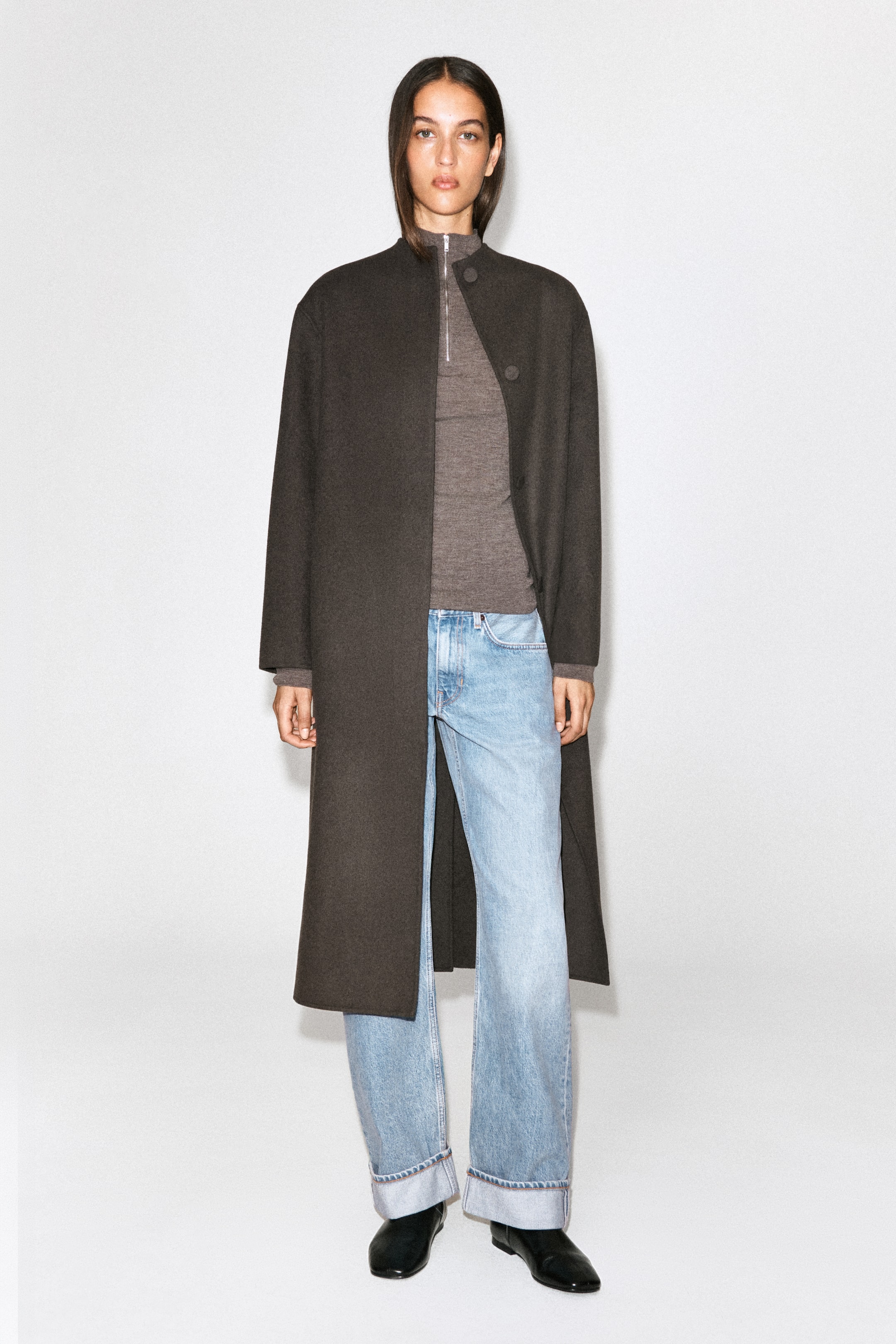 Se større billede: Lyseblå Folde-jeans med buede ben og opsmøg, der har en afslappet silhuet og brede opfoldede manchetter, bæres af modellen. Jeansene styles med en lang, mørkebrun frakke og sorte læderstøvler.
