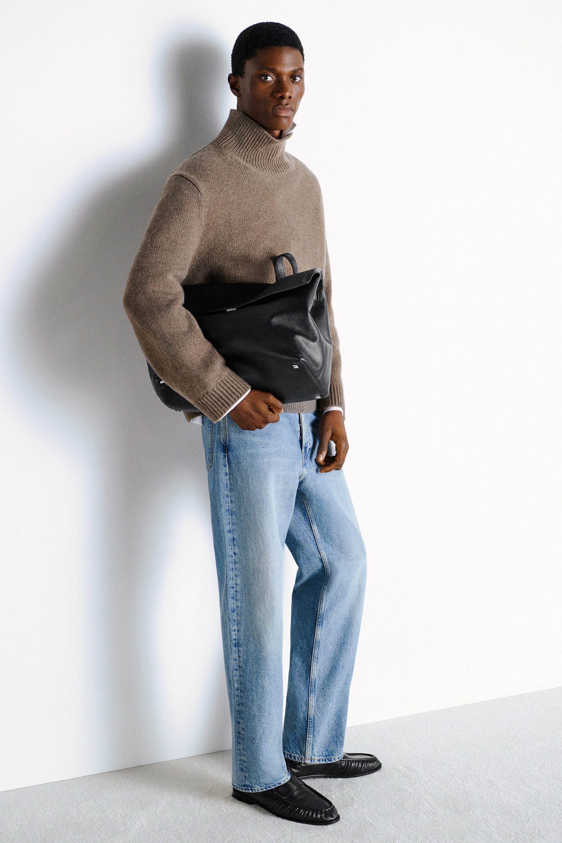 Ein taupebrauner Kaschmir-Pullover wird aus einer dreiviertel Frontansicht gezeigt und verfügt über eine lockere Passform, einen hohen gerippten Stehkragen und abgerundete Ärmel mit gerippten Bündchen. Das Model trägt den Strickpullover zusammen mit hellblauen Jeans und hält eine grosse schwarze Ledertasche. Ein taupebrauner Kaschmir-Pullover wird aus einer dreiviertel Frontansicht gezeigt und verfügt über eine lockere Passform, einen hohen gerippten Stehkragen und abgerundete Ärmel mit gerippten Bündchen. Das Model trägt den Strickpullover zusammen mit hellblauen Jeans und hält eine grosse schwarze Ledertasche.