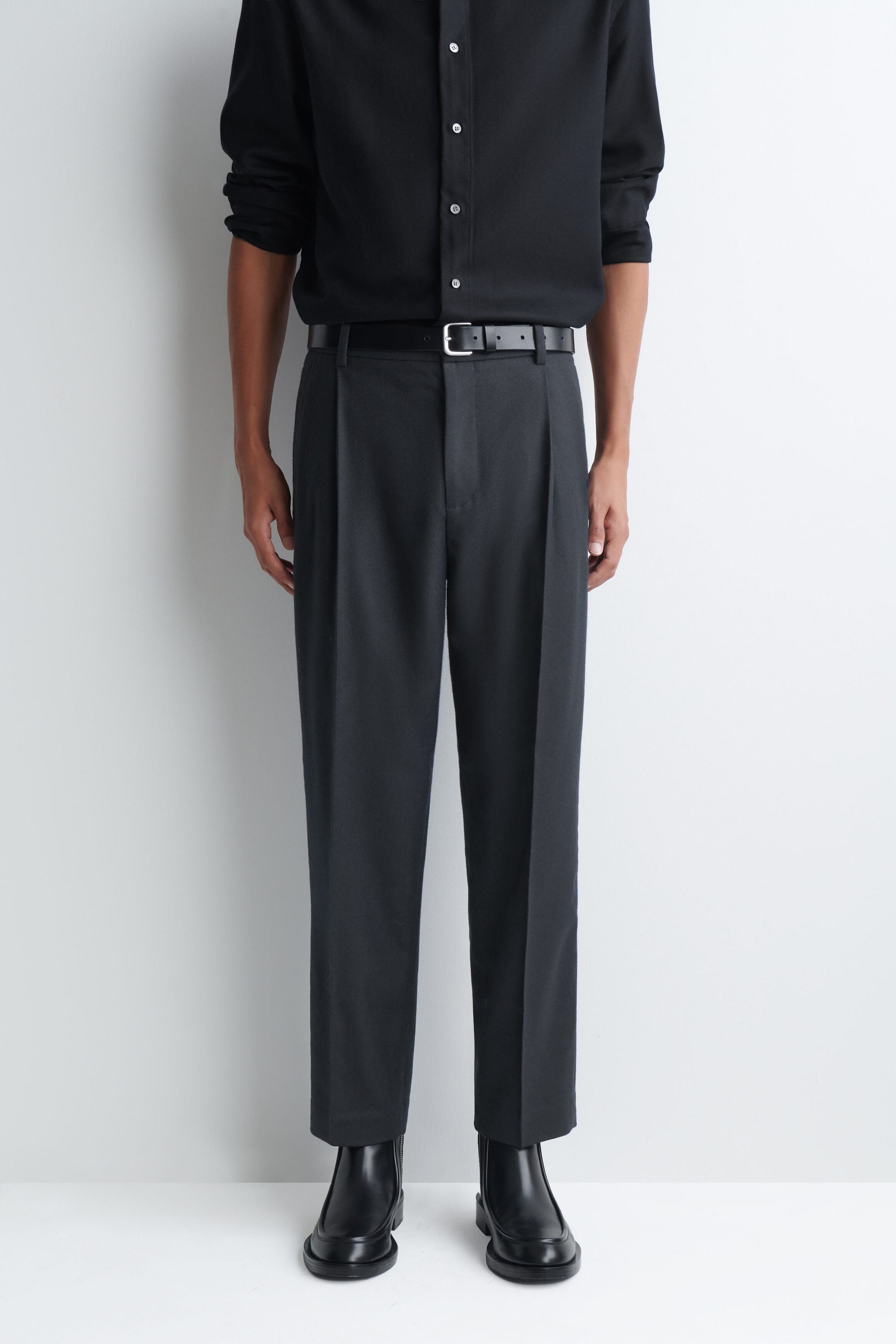 The model is wearing dark gray pleated trousers, featuring a straight-leg silhouette with sharp pressed creases down the front. The trousers are styled with a tucked-in black button-down shirt, a black leather belt with a silver buckle, and black leather boots. The model is wearing dark gray pleated trousers, featuring a straight-leg silhouette with sharp pressed creases down the front. The trousers are styled with a tucked-in black button-down shirt, a black leather belt with a silver buckle, and black leather boots.
