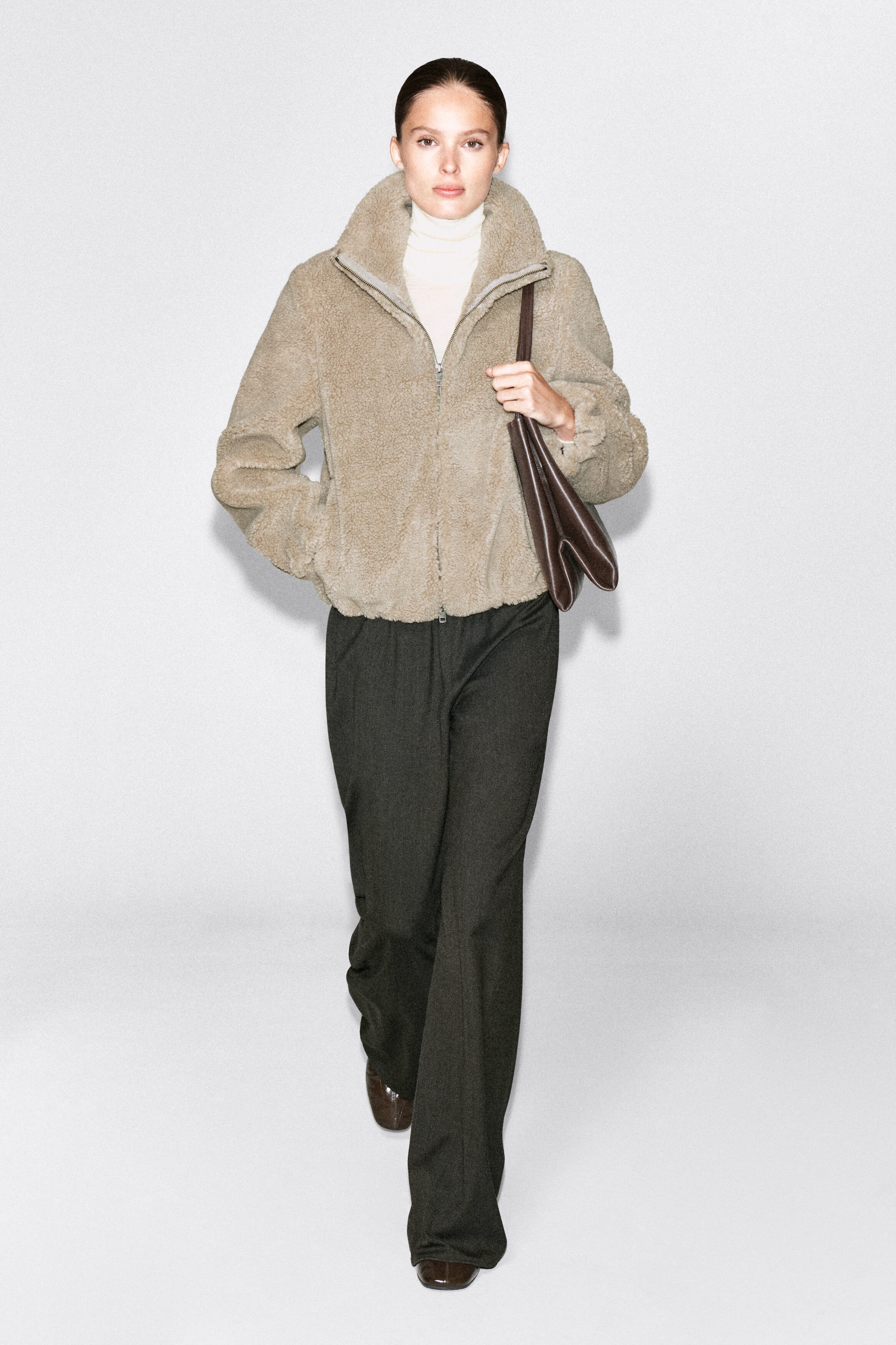 The model is wearing a beige funnel-neck teddy jacket, walking with one hand resting in the side pocket. The textured fleece jacket is layered over a cream turtleneck sweater and styled with dark wide-leg trousers and a brown leather shoulder bag.