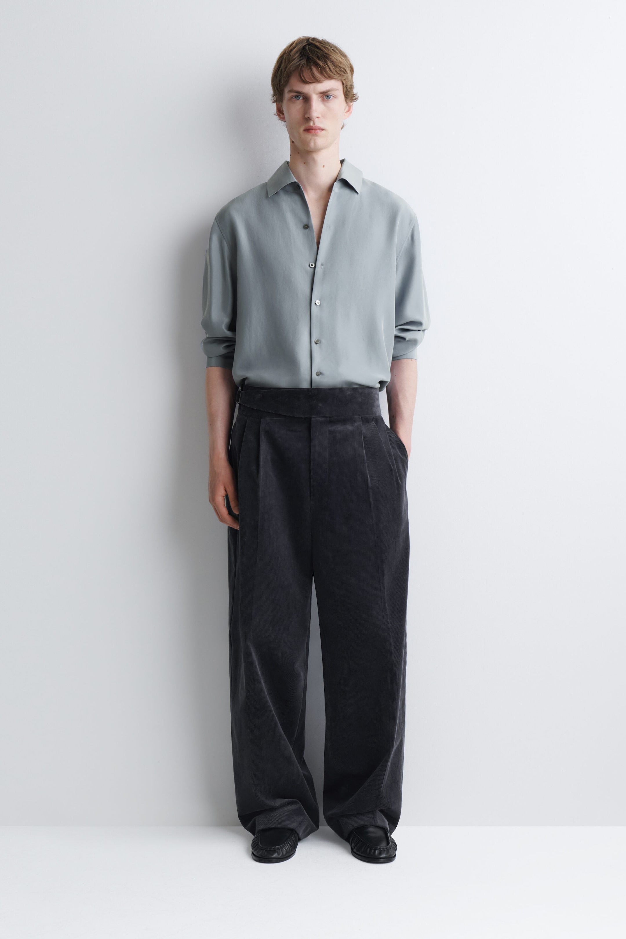The model is wearing a relaxed silk shirt in a light grey-green hue, featuring a pointed collar, button fastening and long sleeves. The shirt is tucked into high-waisted, wide-leg dark trousers to emphasise its fluid drape. The model is wearing a relaxed silk shirt in a light grey-green hue, featuring a pointed collar, button fastening and long sleeves. The shirt is tucked into high-waisted, wide-leg dark trousers to emphasise its fluid drape.