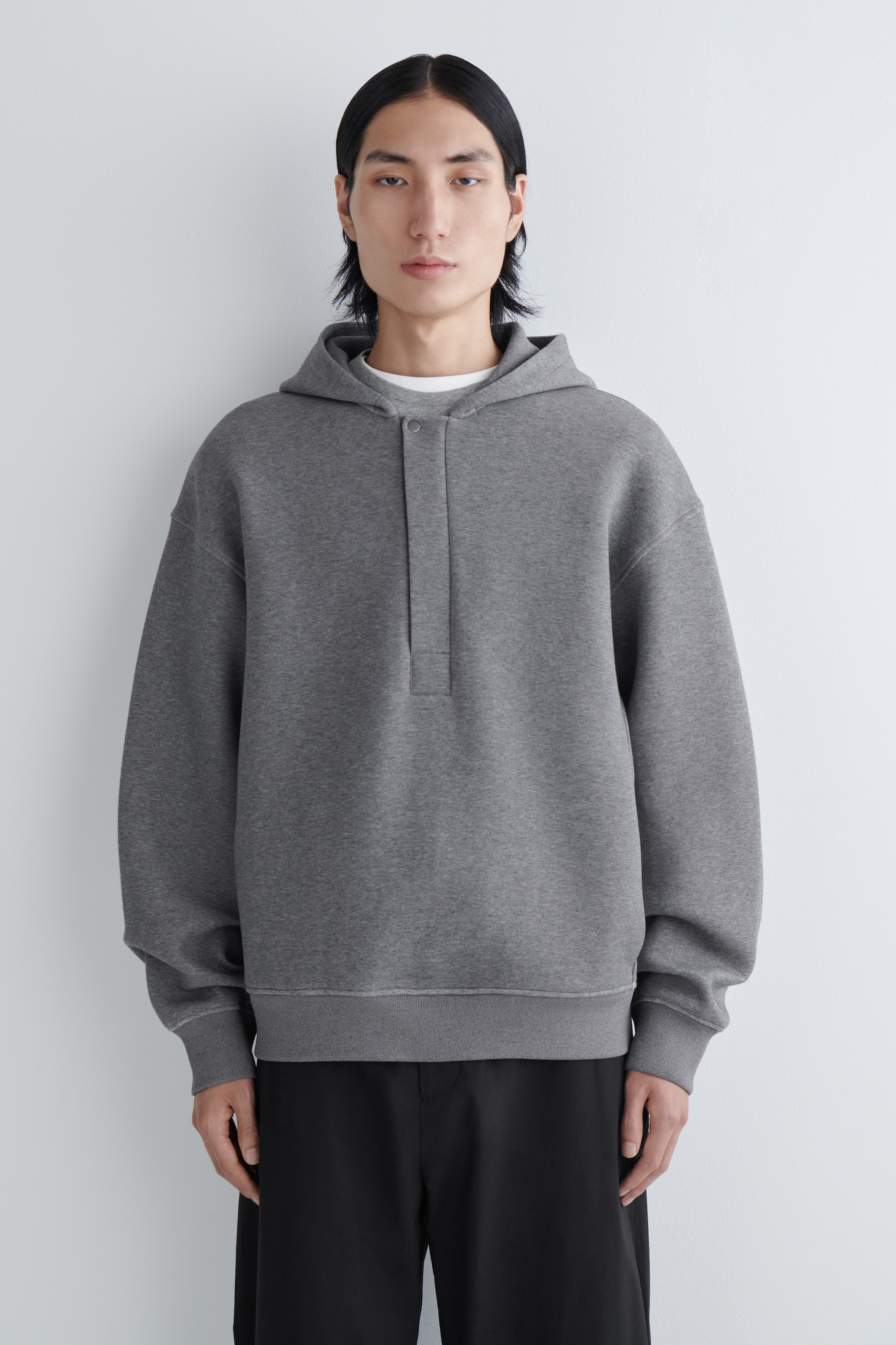 Zobrazit větší obrázek: Šedá mikina s kapucí je prezentována zepředu, má volný střih a kapuci. Je zdobena krátkou zapínací légou u výstřihu, žebrovanými manžetami na dlouhých rukávech a žebrovaným lemem, pod kterou je navrstvené bílé tričko s kulatým výstřihem.