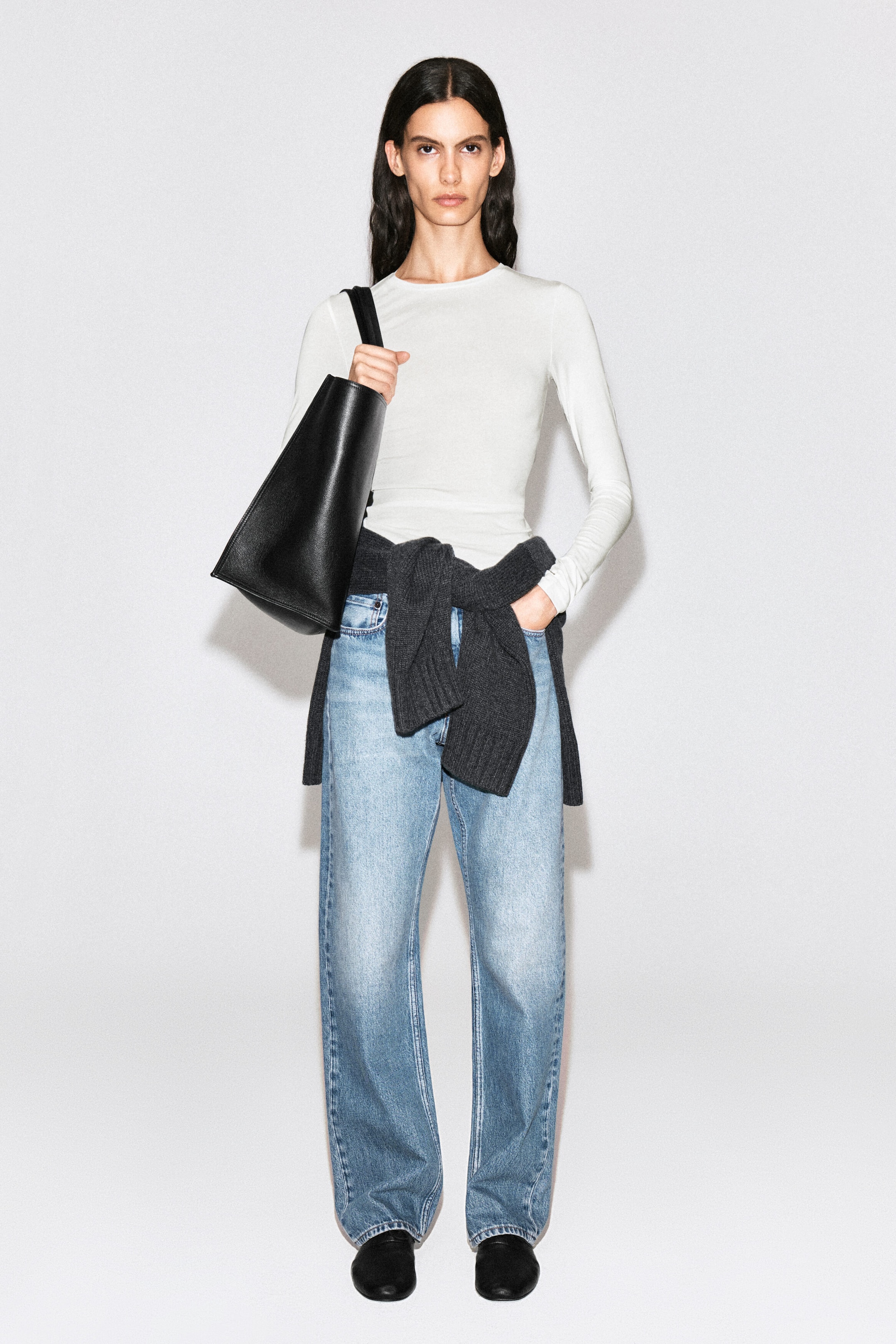 A black leather tote bag with a single shoulder strap is being carried over the model's shoulder, with the strap held in one hand. The bag is styled with a white long-sleeve top, light-wash wide-leg jeans, and a dark gray sweater tied around the waist.