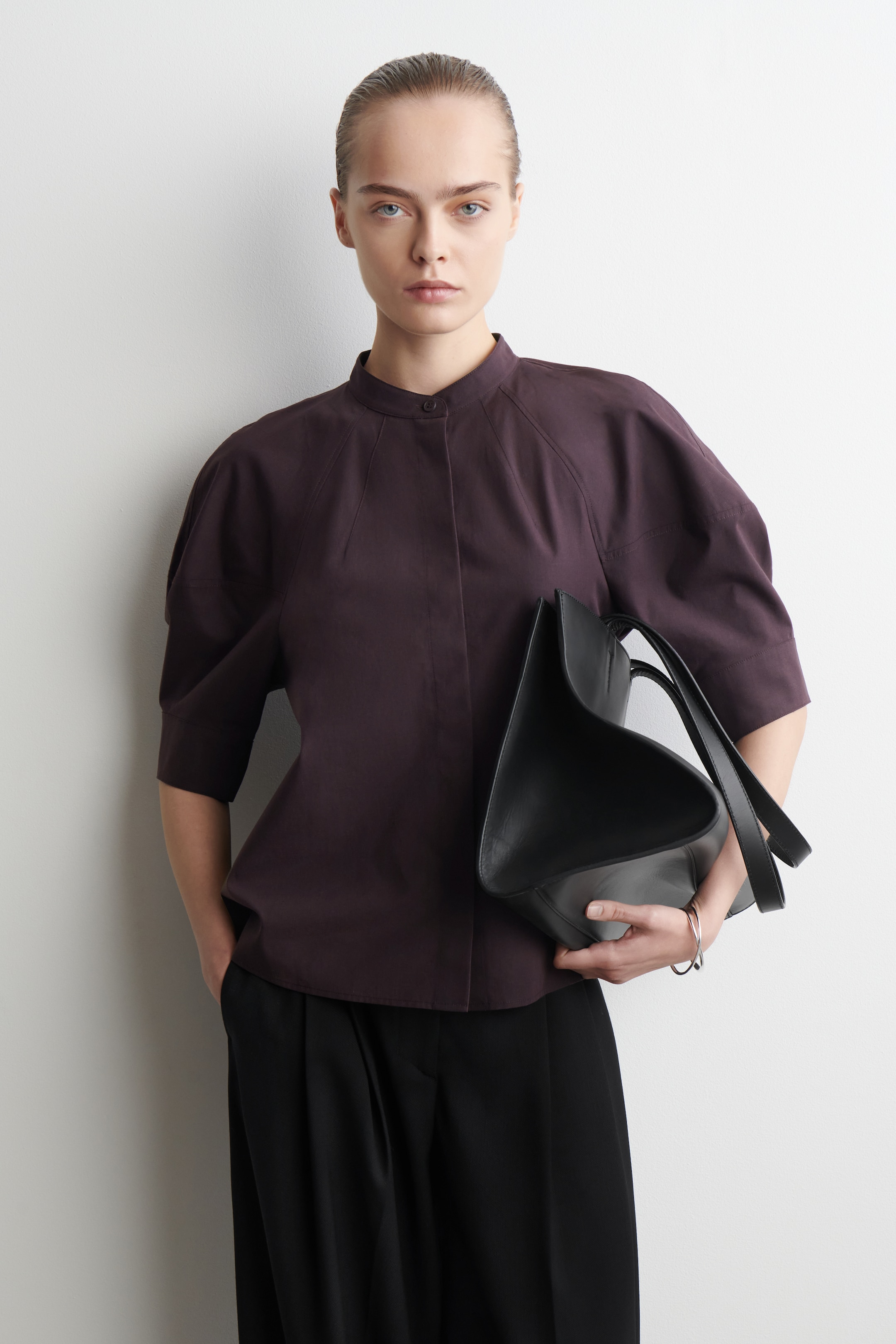 Visualizza immagine più grande: Una blusa di colore prugna scuro viene presentata, caratterizzata da un colletto a fascia con chiusura a bottone singolo, cuciture raglan prominenti e maniche voluminose lunghe fino al gomito. Viene indossata con pantaloni neri a gamba larga ed è abbinata a una borsa tote scultorea nera tenuta in primo piano.