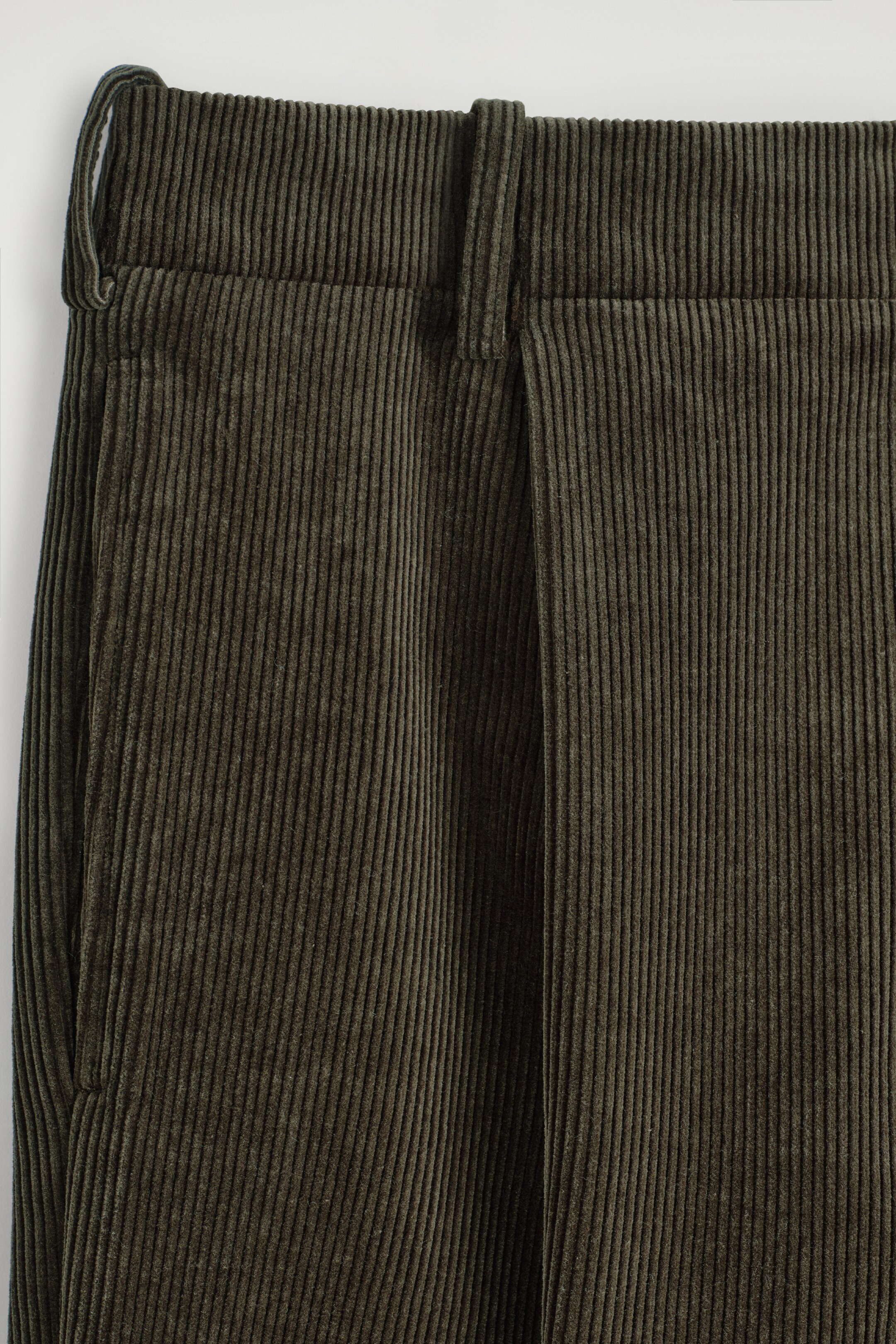Agrandir l'image: Un pantalon en velours côtelé vert foncé est présenté de face, affichant une texture finement côtelée. La ceinture plate est dotée de passants de ceinture visibles, et un seul pli s'étend le long de la jambe avant droite depuis la ceinture, avec une ouverture de poche latérale apparaissant sur la gauche.