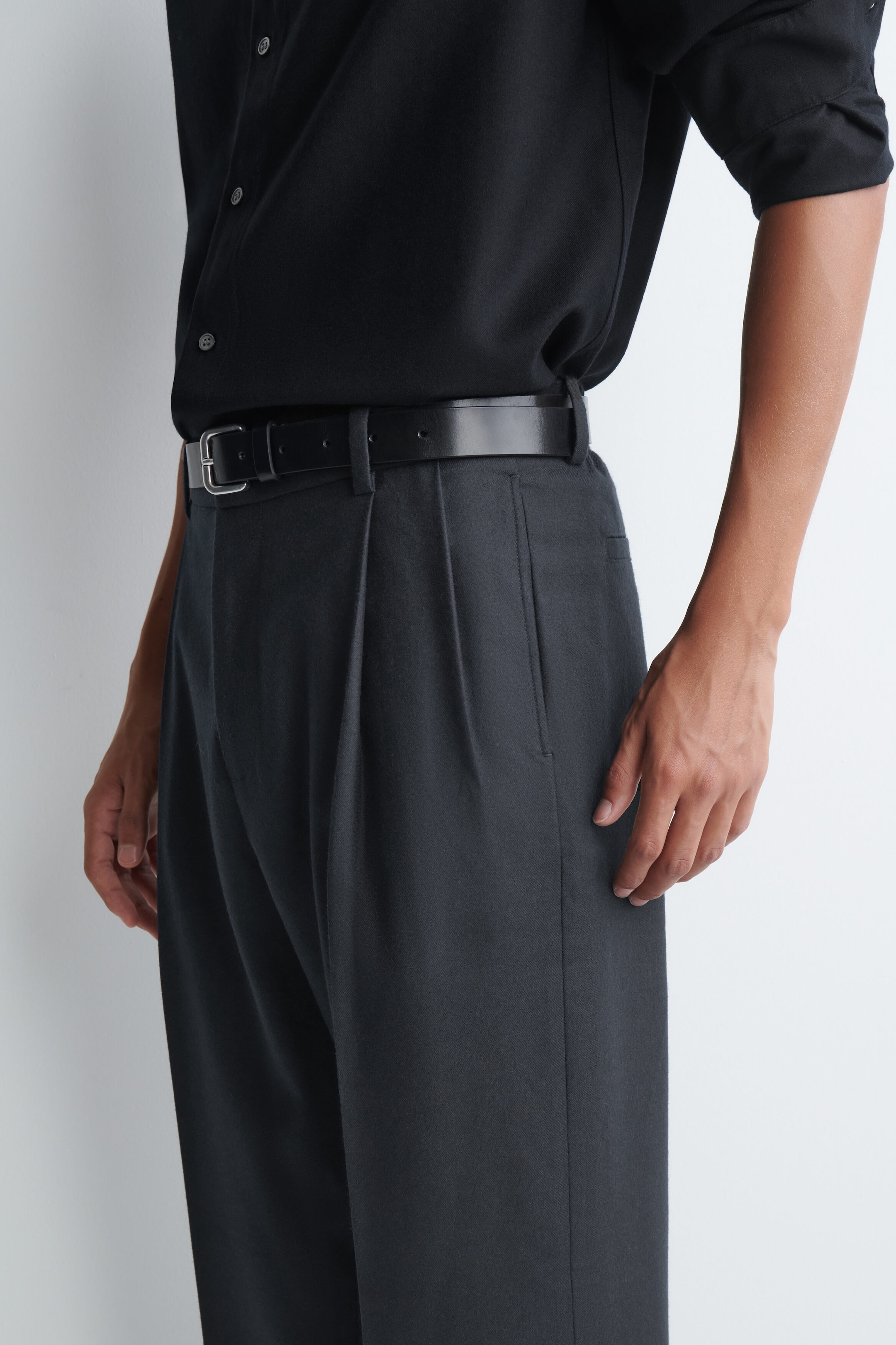 The model is wearing dark gray pleated trousers, styled with a black leather belt and a tucked-in black button-up shirt. The trousers feature front pleats and side pockets and sit high on the waist. The model is wearing dark gray pleated trousers, styled with a black leather belt and a tucked-in black button-up shirt. The trousers feature front pleats and side pockets and sit high on the waist.