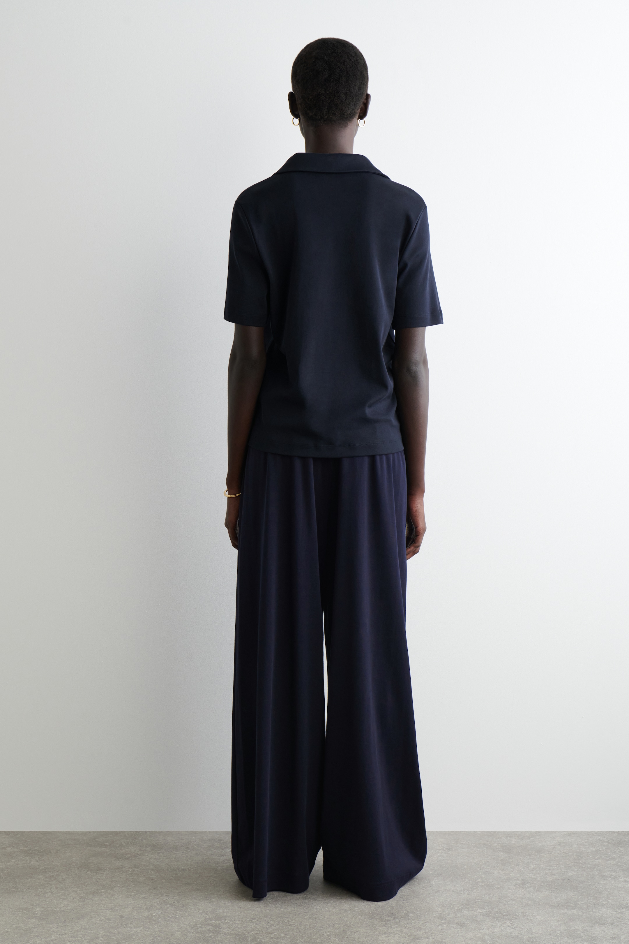 Agrandir l'image: Un polo à manches courtes bleu foncé et un pantalon large assorti sont portés, vus de dos. Le polo est stylé avec un col classique et une coupe décontractée, tandis que le pantalon présente une silhouette longue et large.