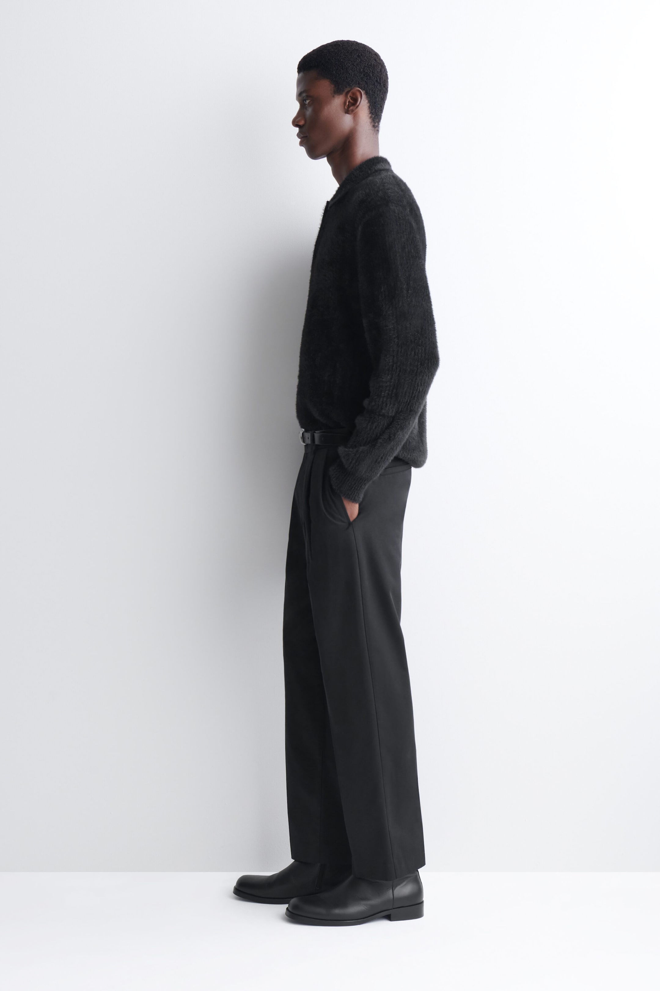 A black knitted polo shirt with a highly textured, brushed finish is worn by the model, who is standing in profile. The long-sleeved shirt is styled tucked into tailored black trousers with a black leather belt and paired with black leather boots. A black knitted polo shirt with a highly textured, brushed finish is worn by the model, who is standing in profile. The long-sleeved shirt is styled tucked into tailored black trousers with a black leather belt and paired with black leather boots.
