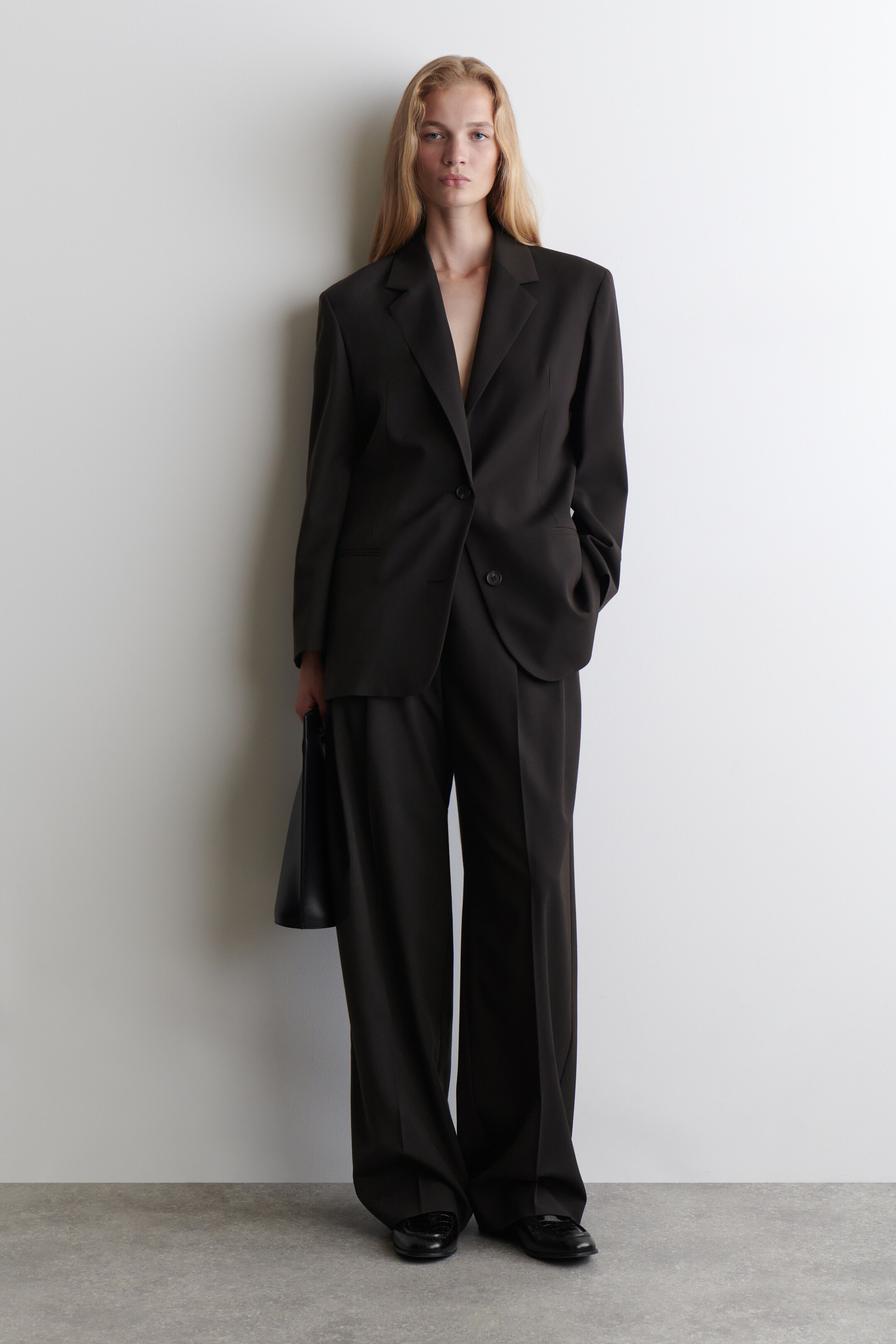 Agrandir l'image: Un costume en charbon foncé est présenté avec une coupe décontractée et surdimensionnée. Le blazer à simple boutonnage présente des revers crantés et une fermeture à un bouton, tandis que le pantalon assorti affiche une silhouette à jambes larges.