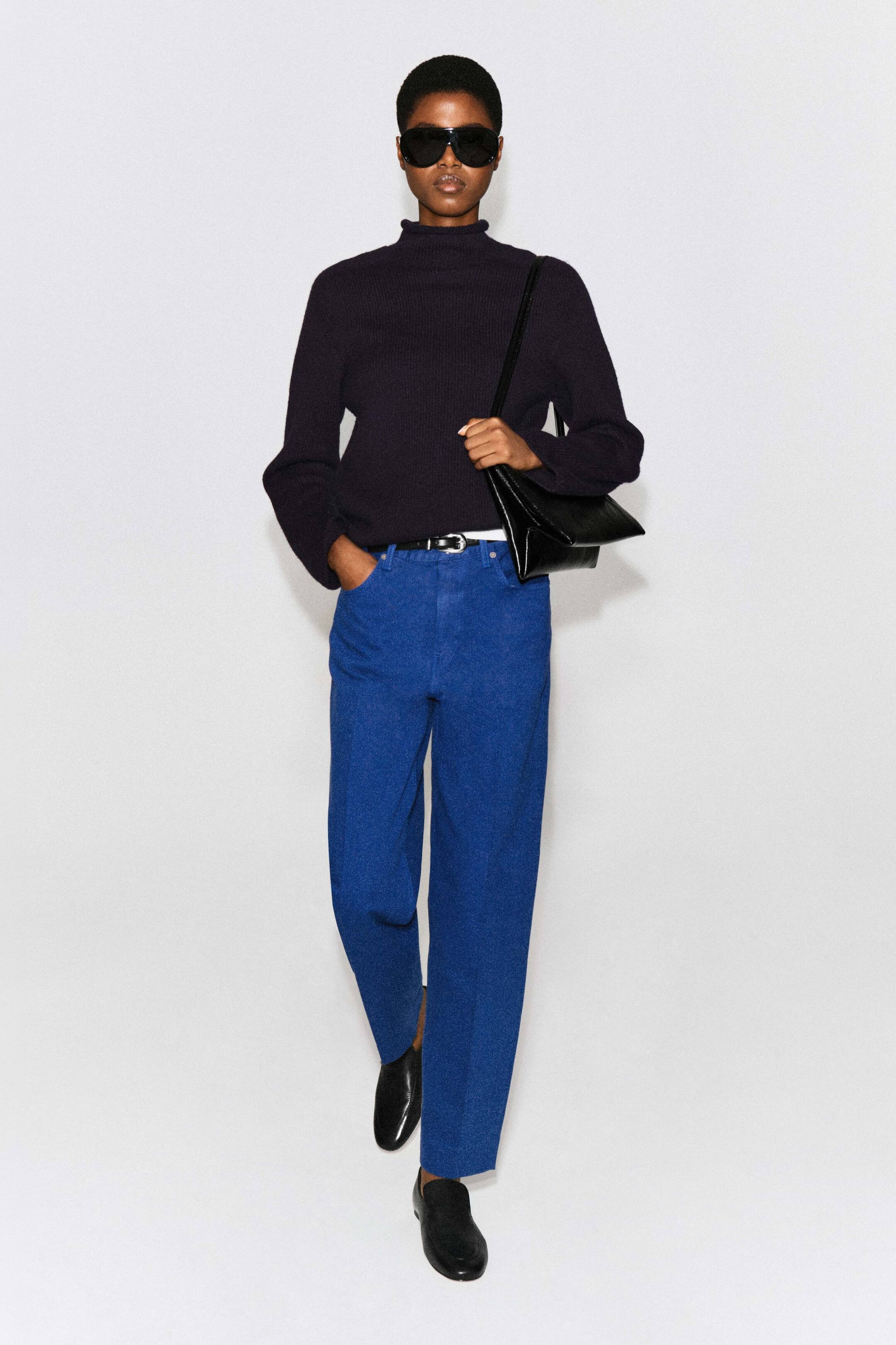 Agrandir l'image: Le jean tapered Umbra dans une teinte bleu vif est porté par le mannequin, avec une taille haute, une jambe fuselée décontractée et des plis centraux marqués. Le jean est porté avec une ceinture noire, un pull foncé à col montant rentré et des chaussures noires, tandis que le mannequin repose une main dans une poche avant.