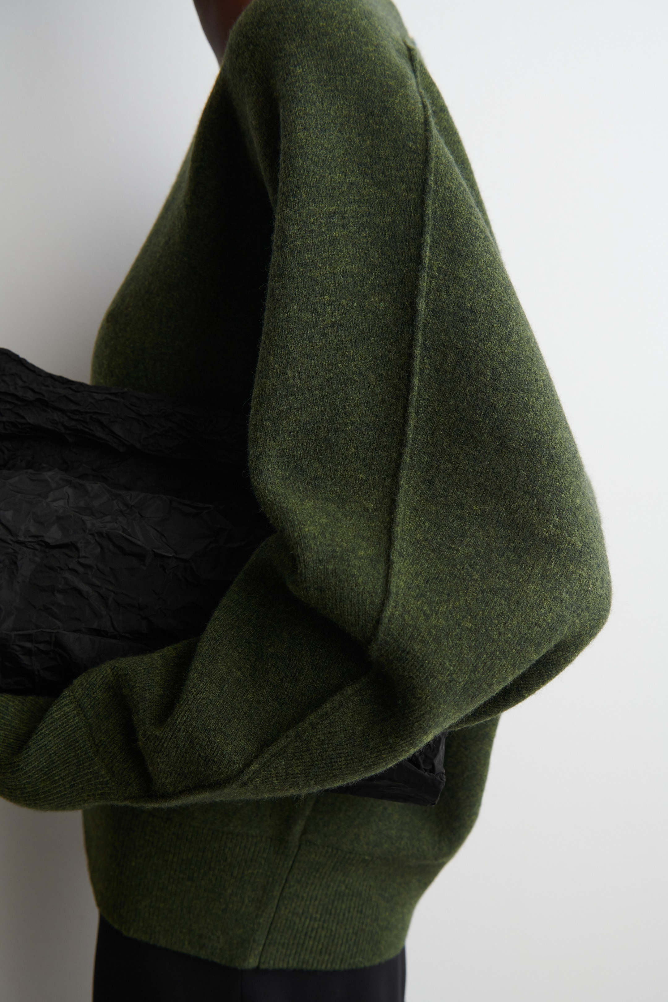 Grösseres Bild ansehen: Ein grüner Mélange-Strickpullover wird aus einer Seitenansicht präsentiert, mit einer lockeren Passform und langen, voluminösen Ärmeln, die durch eine markante vertikale Naht und einen gerippten Saum hervorgehoben werden. Ein schwarzes, zerknittertes, strukturiertes Objekt wird von der Trägerin gehalten und ist teilweise unter dem Ärmel des Pullovers sichtbar, was den großzügigen Fall des Kleidungsstücks betont.
