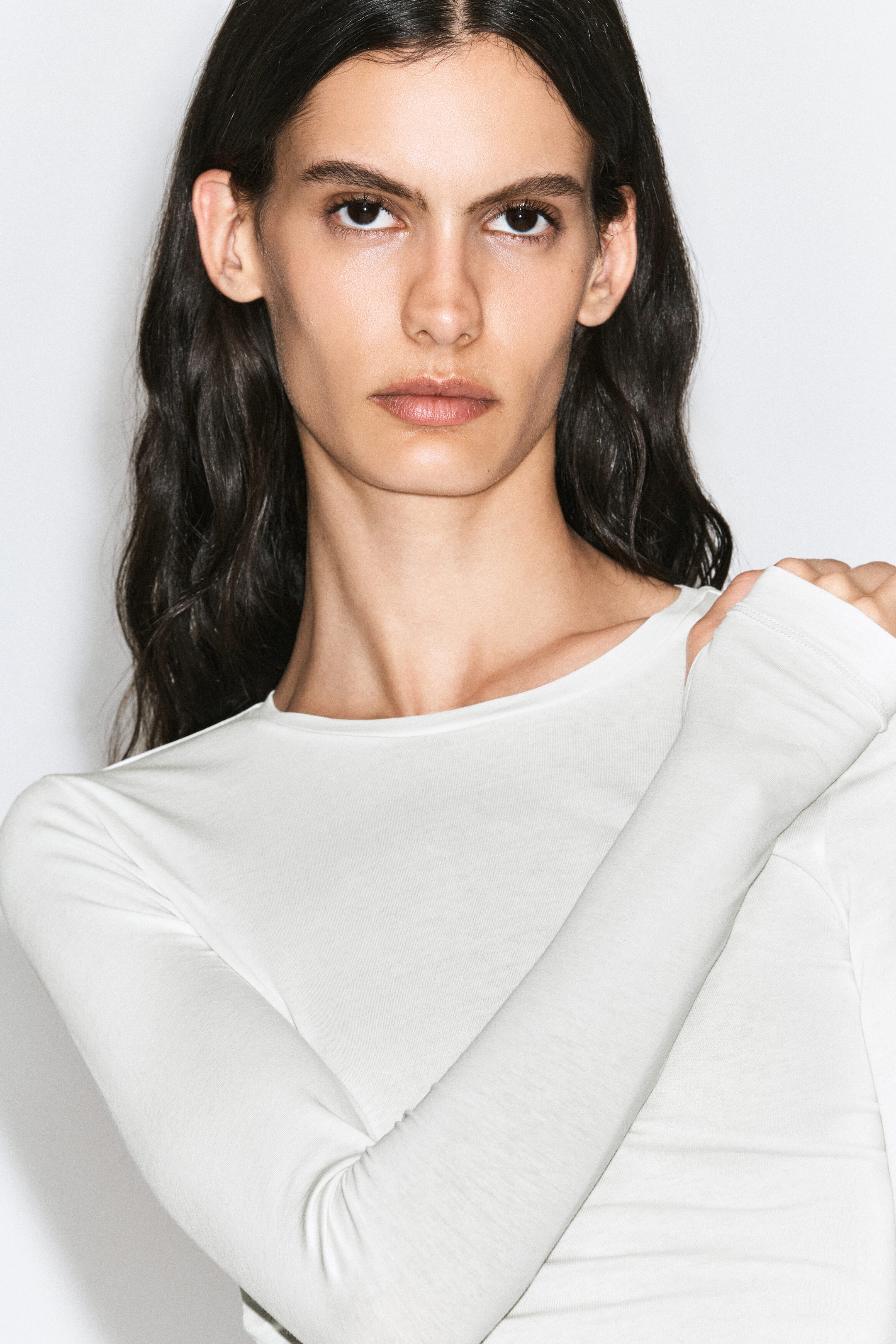 A white, long-sleeved crewneck top is being worn by the model. The model is holding her opposite shoulder, showcasing the fitted sleeve and lightweight jersey material of the top.