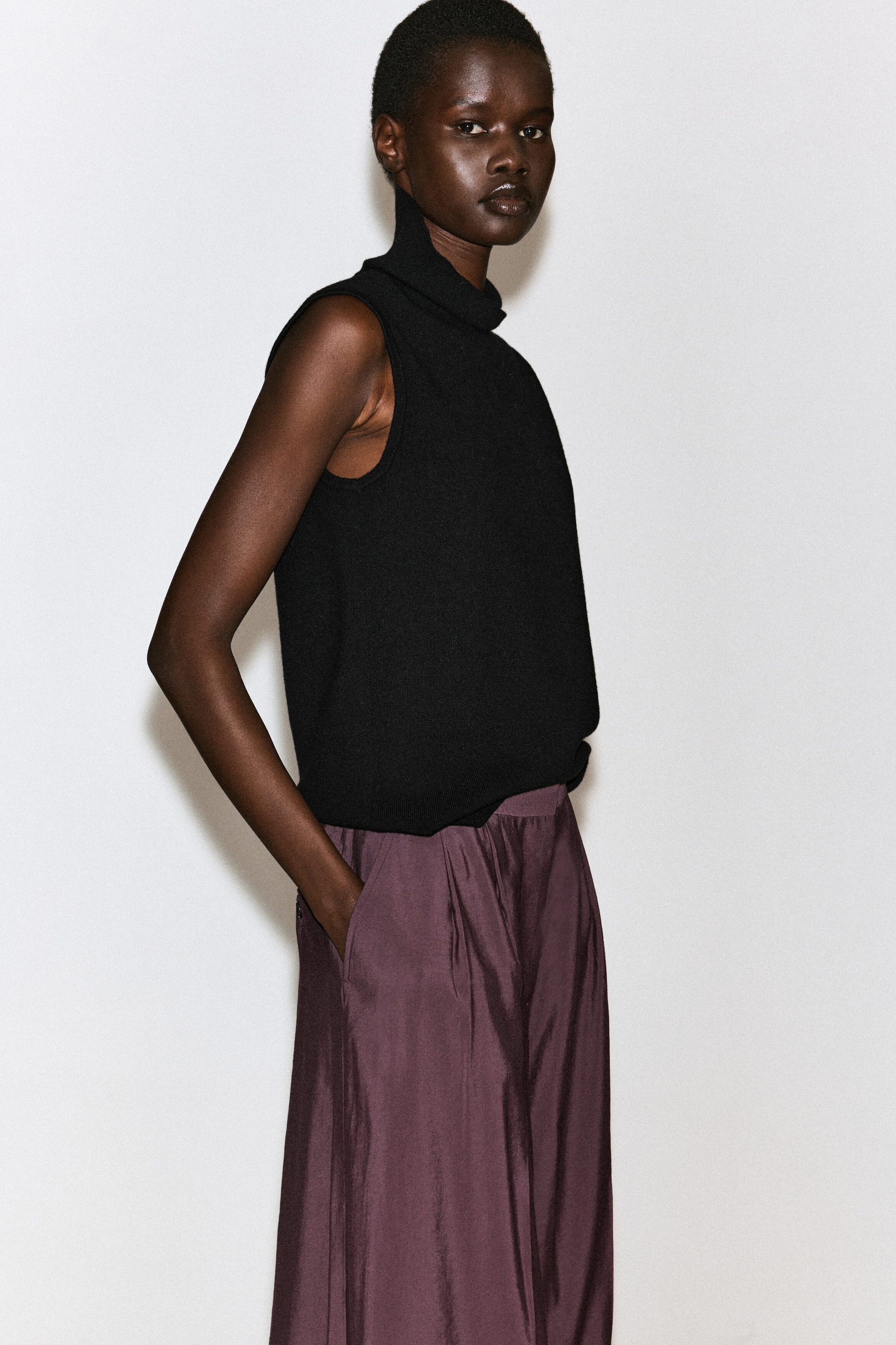 Eine plissierte Wide-Leg-Hose in dunklem Pflaumenton wird von dem Model getragen, das mit einer Hand in der Seitentasche steht. Die Hose hat einen leichten Glanz und eine Plissierung an der Taille und wird mit einem schwarzen ärmellosen Rollkragenoberteil kombiniert. Eine plissierte Wide-Leg-Hose in dunklem Pflaumenton wird von dem Model getragen, das mit einer Hand in der Seitentasche steht. Die Hose hat einen leichten Glanz und eine Plissierung an der Taille und wird mit einem schwarzen ärmellosen Rollkragenoberteil kombiniert.