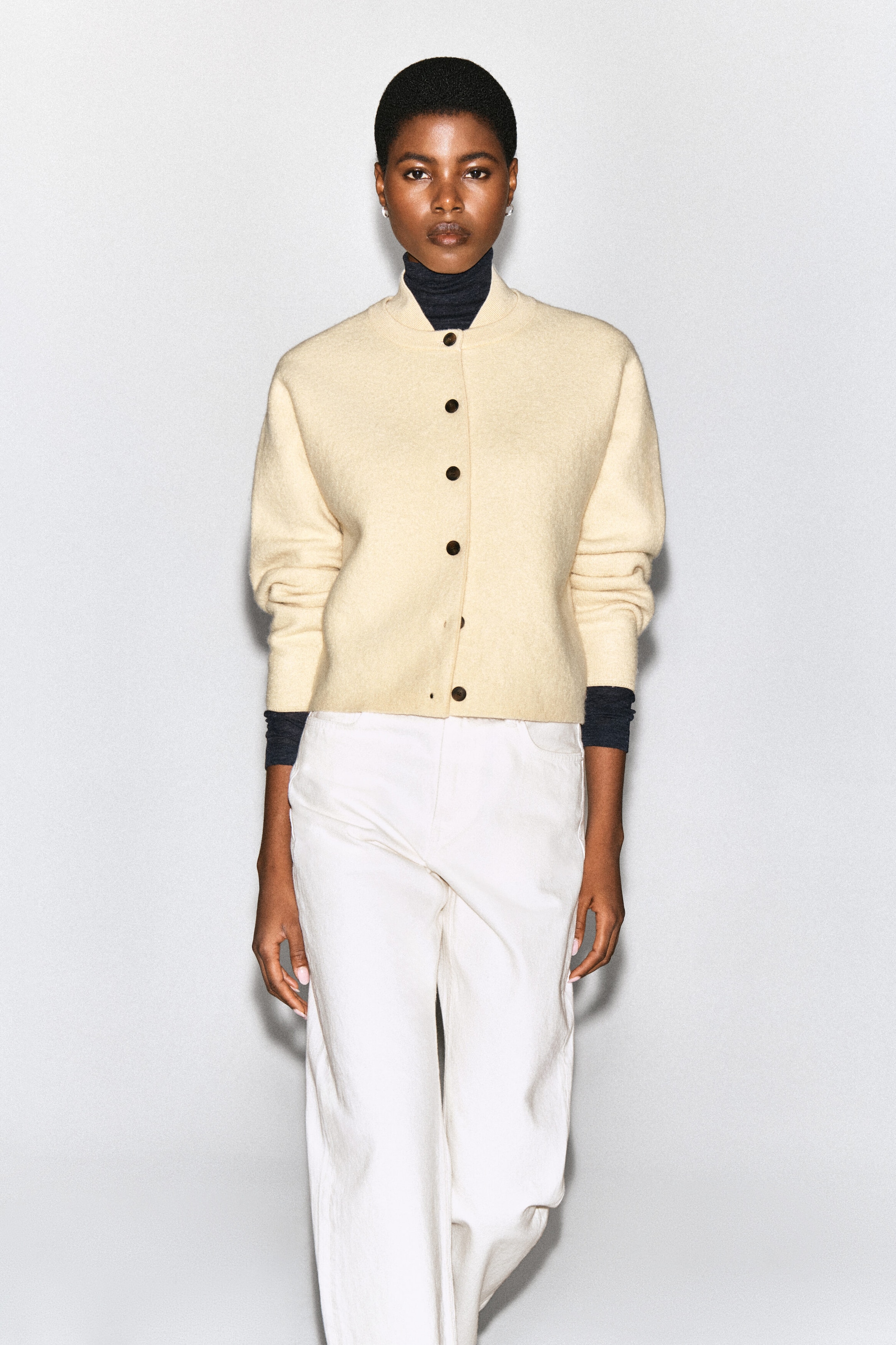 Le mannequin porte un cardigan en laine bouillie de couleur crème, de forme arrondie, avec un col de style bomber et des boutons foncés à l’avant. Le cardigan est superposé à un col roulé gris foncé et agencé avec un pantalon blanc. Le mannequin porte un cardigan en laine bouillie de couleur crème, de forme arrondie, avec un col de style bomber et des boutons foncés à l’avant. Le cardigan est superposé à un col roulé gris foncé et agencé avec un pantalon blanc.