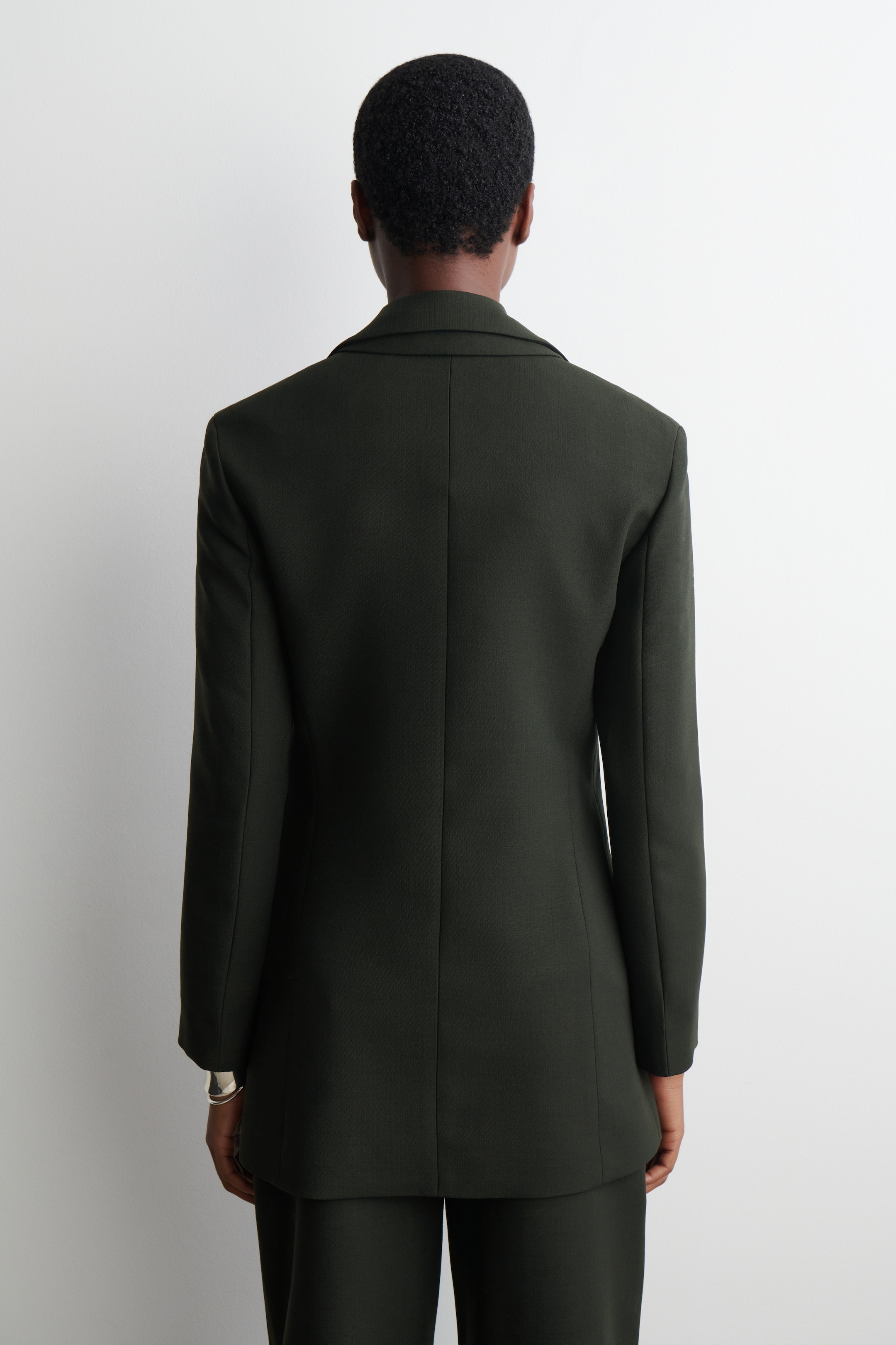 Grösseres Bild ansehen: Ein dunkelgrüner Blazer wird von hinten präsentiert und hebt seine maßgeschneiderte Passform hervor. Er verfügt über einen strukturierten Kragen und vertikale Nähte, mit langen Ärmeln und einem Saum, der über passenden dunkelgrünen Hosen verläuft.