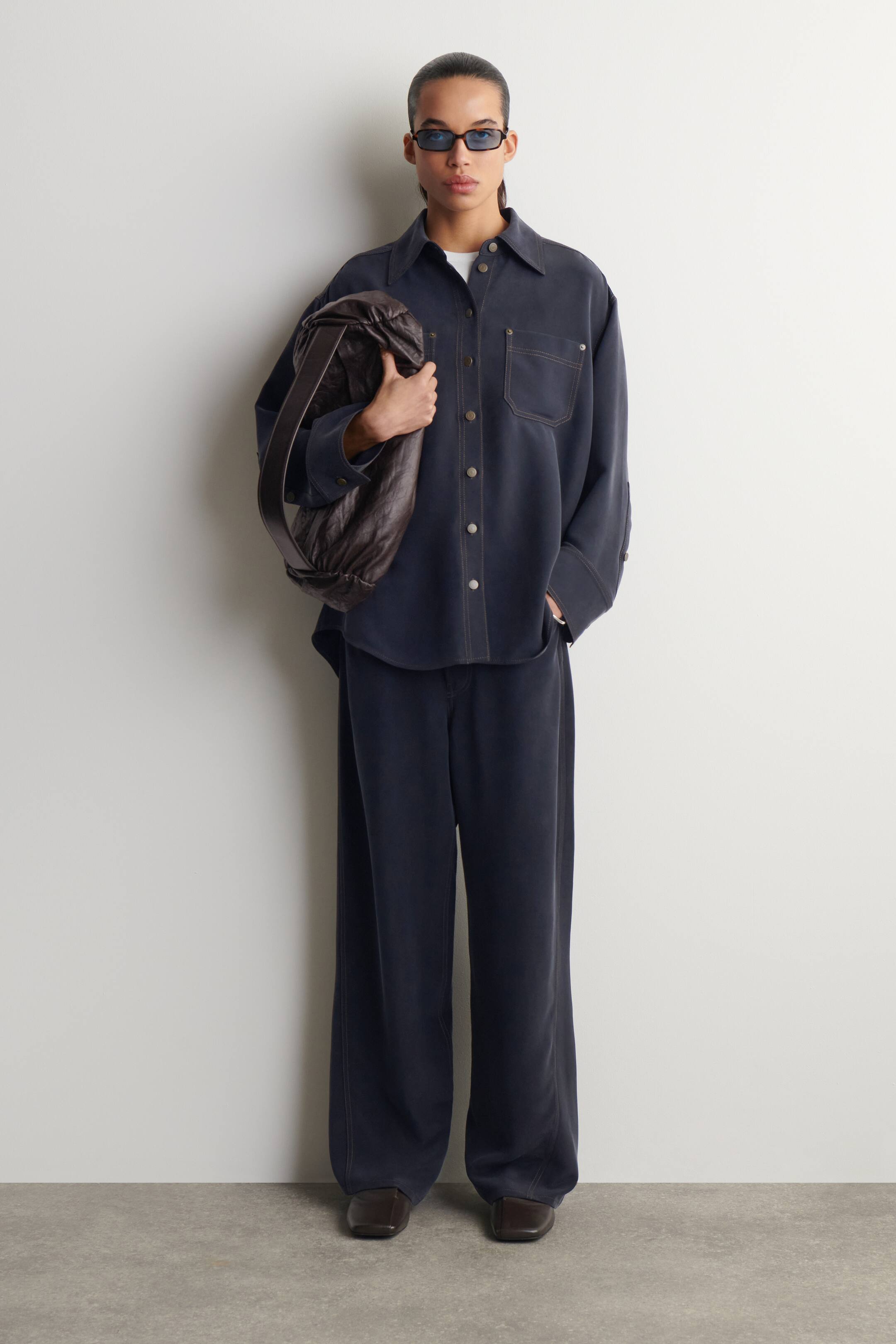 Agrandir l'image: Une chemise bleu foncé à manches longues et à boutons, ainsi qu'un pantalon large assorti, sont présentés de face. La chemise est dotée d'un col en pointe, de boutons de couleur claire, de deux poches poitrine à rabat et de surpiqûres contrastées, et est portée par-dessus un t-shirt blanc. Le pantalon à coupe décontractée affiche également des surpiqûres contrastées.