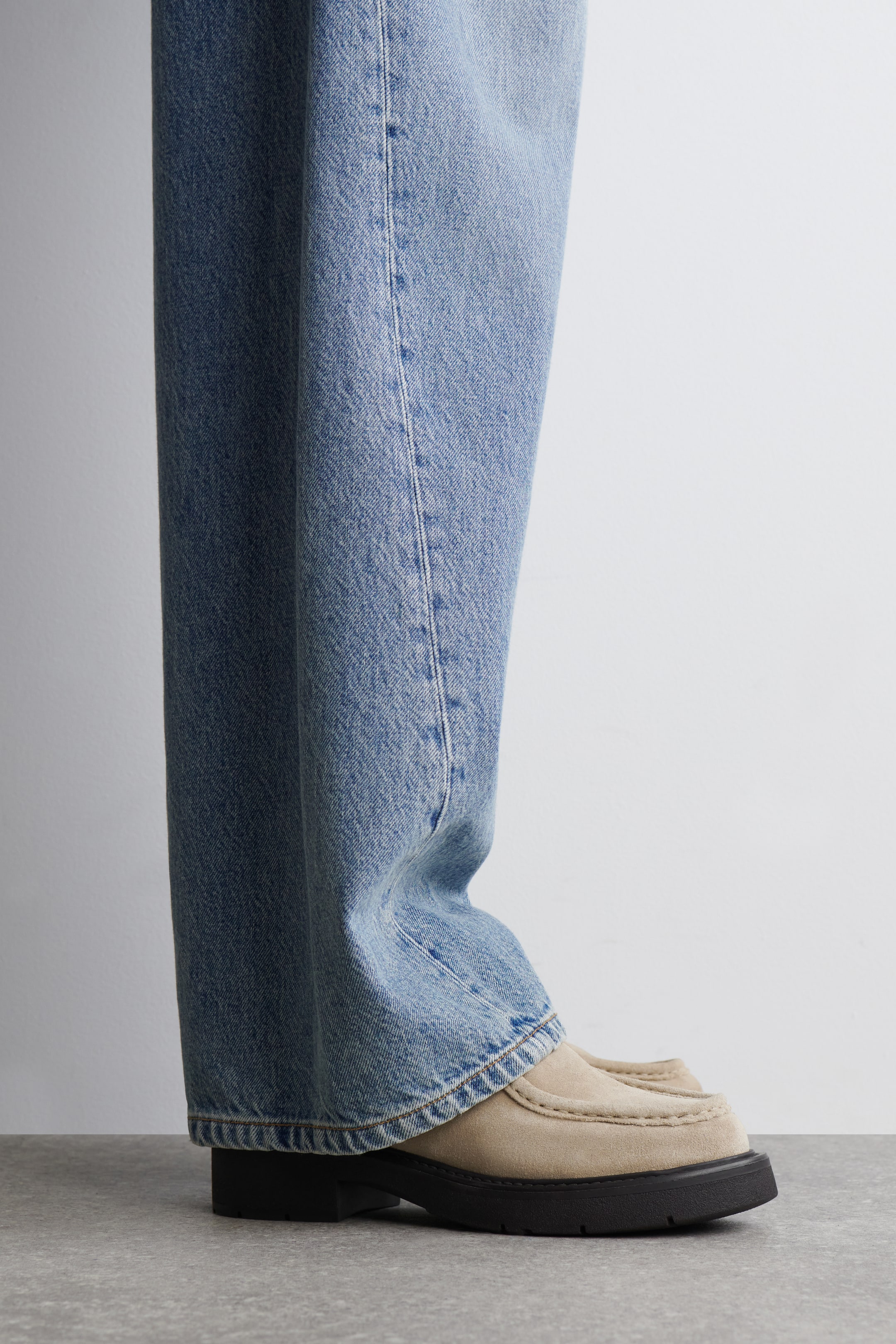 Agrandir l'image: Une paire de jeans en denim bleu clair à jambes larges s'accumule sur le dessus de mocassins en daim beige. Les mocassins présentent des coutures de style mocassin proéminentes sur l'orteil et une semelle épaisse en caoutchouc noir, visible sous un angle de côté.