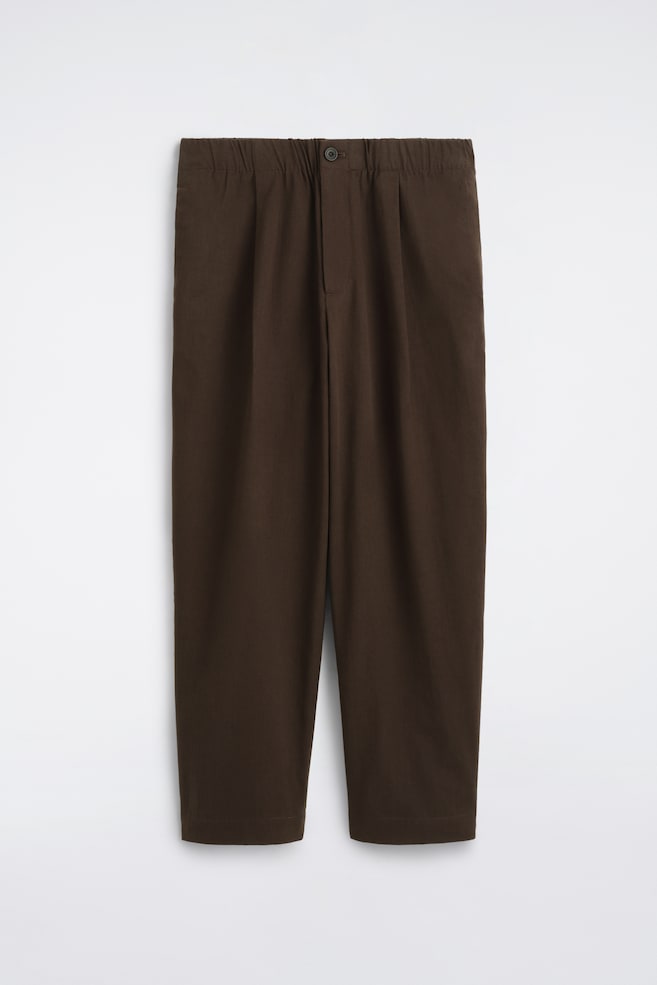 Diese dunkelbraunen, plissierten Barrel-Leg-Hosen in Relaxed Fit sind von vorne zu sehen und verfügen über einen elastischen Bund mit mittigem Knopfverschluss. Das Design zeigt eine entspannte, leicht zulaufende Beinsilhouette mit Falten vorne, die für strukturiertes Volumen sorgen. - 1