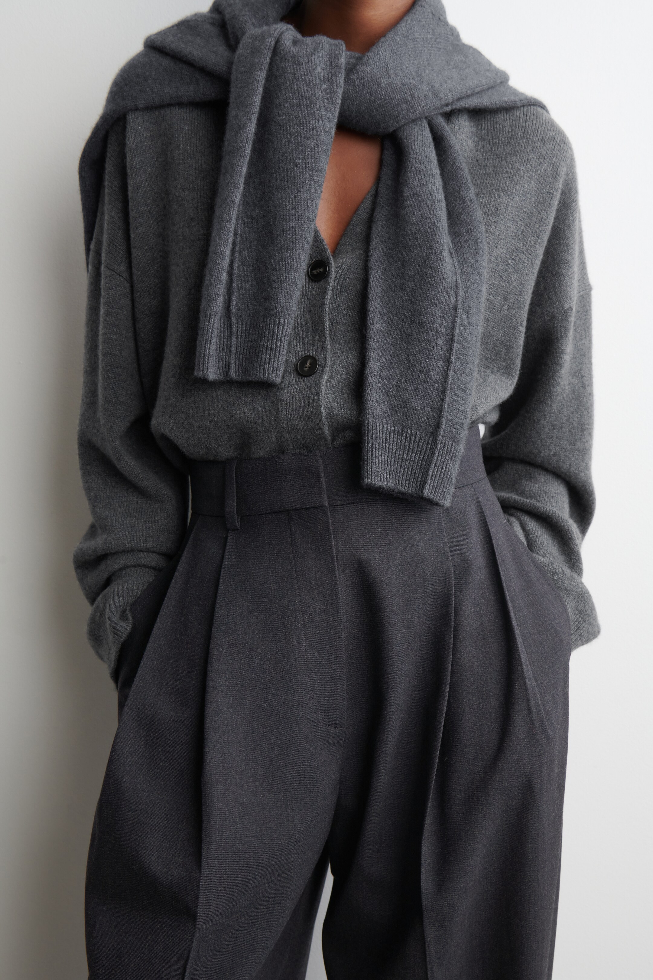 Grösseres Bild ansehen: Ein abgestimmtes Outfit in Dunkelgrau wird aus einem Front-Rechts-Winkel präsentiert. Es zeichnet sich durch eine Strickjacke mit V-Ausschnitt, Knopfverschluss, gerippten Bündchen und Saum, einen passenden Strickschal, der über die Schultern gelegt ist und gerippte Enden hat, sowie hoch taillierte Faltenhosen mit Vorderfalten und Seitentaschen aus.