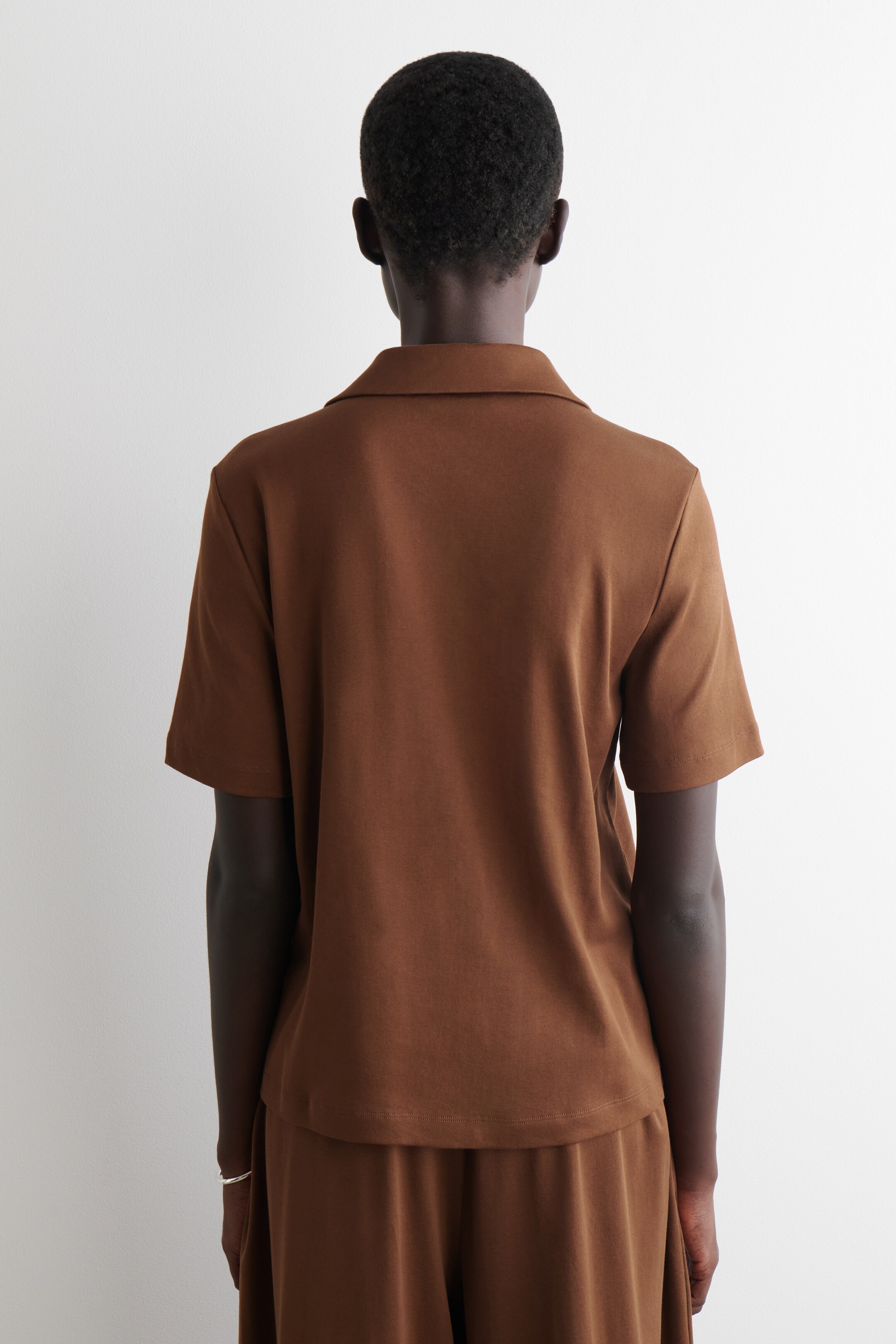 Visualizza immagine più grande: Una polo a maniche corte marrone è esposta dal retro, con un colletto a punta e un orlo dritto, ed è abbinata a pantaloni marroni coordinati.