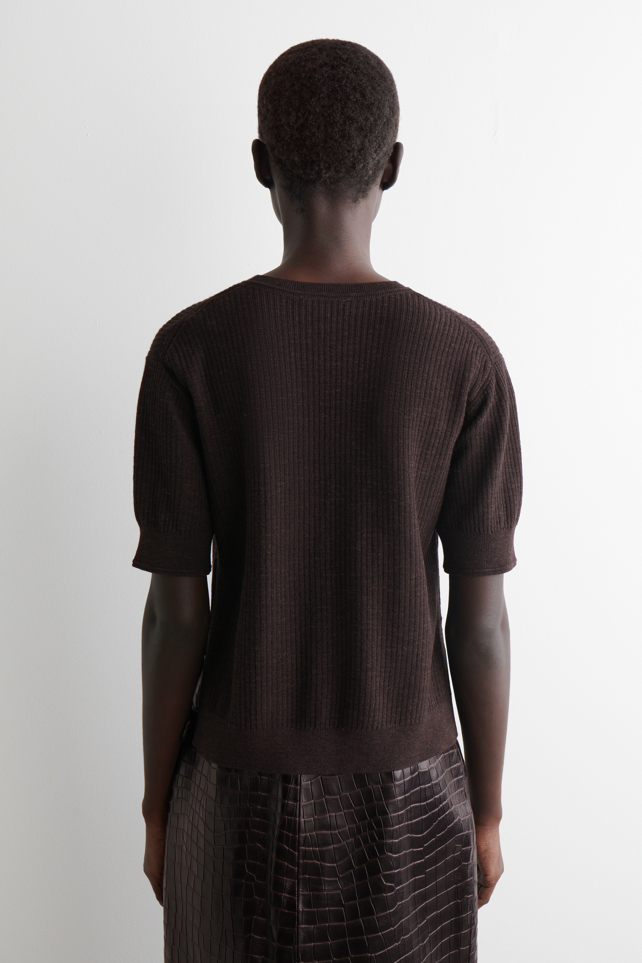 View larger image: A dark brown, ribbed knit top is being worn from the back, featuring short sleeves with ribbed cuffs and a ribbed hem. It is being styled with a dark brown, crocodile-textured skirt.