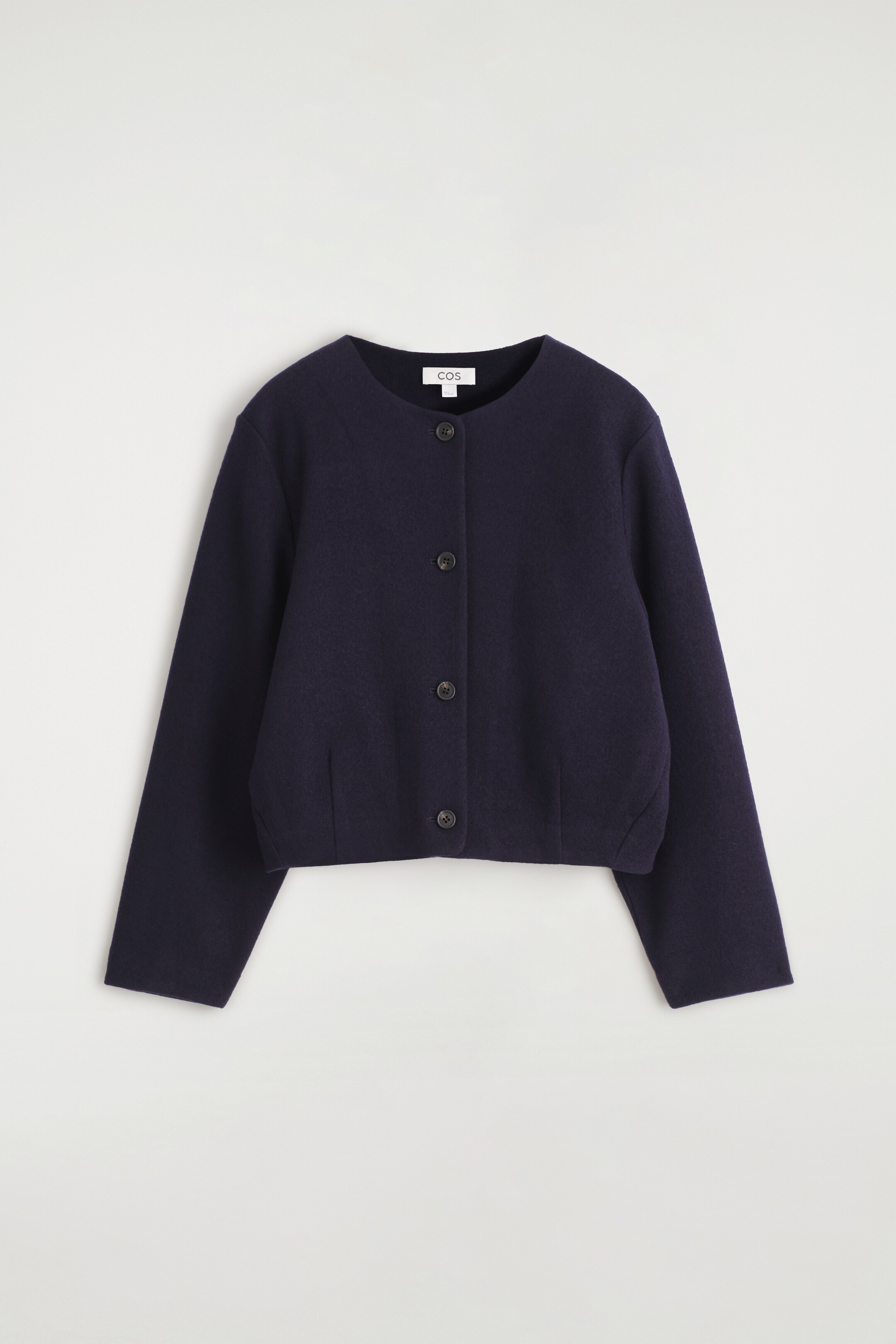 Größeres Bild anzeigen: Eine dunkel marineblaue, kurze Jacke wird von vorne gezeigt, mit einem runden, kragenlosen Ausschnitt und einem vierknöpfigen Verschluss mit dunklen, flachen Knöpfen. Die strukturierte, kastige Jacke zeigt lange, gerade Ärmel und subtile vertikale Nahtdetails in der Nähe ihres Saums.